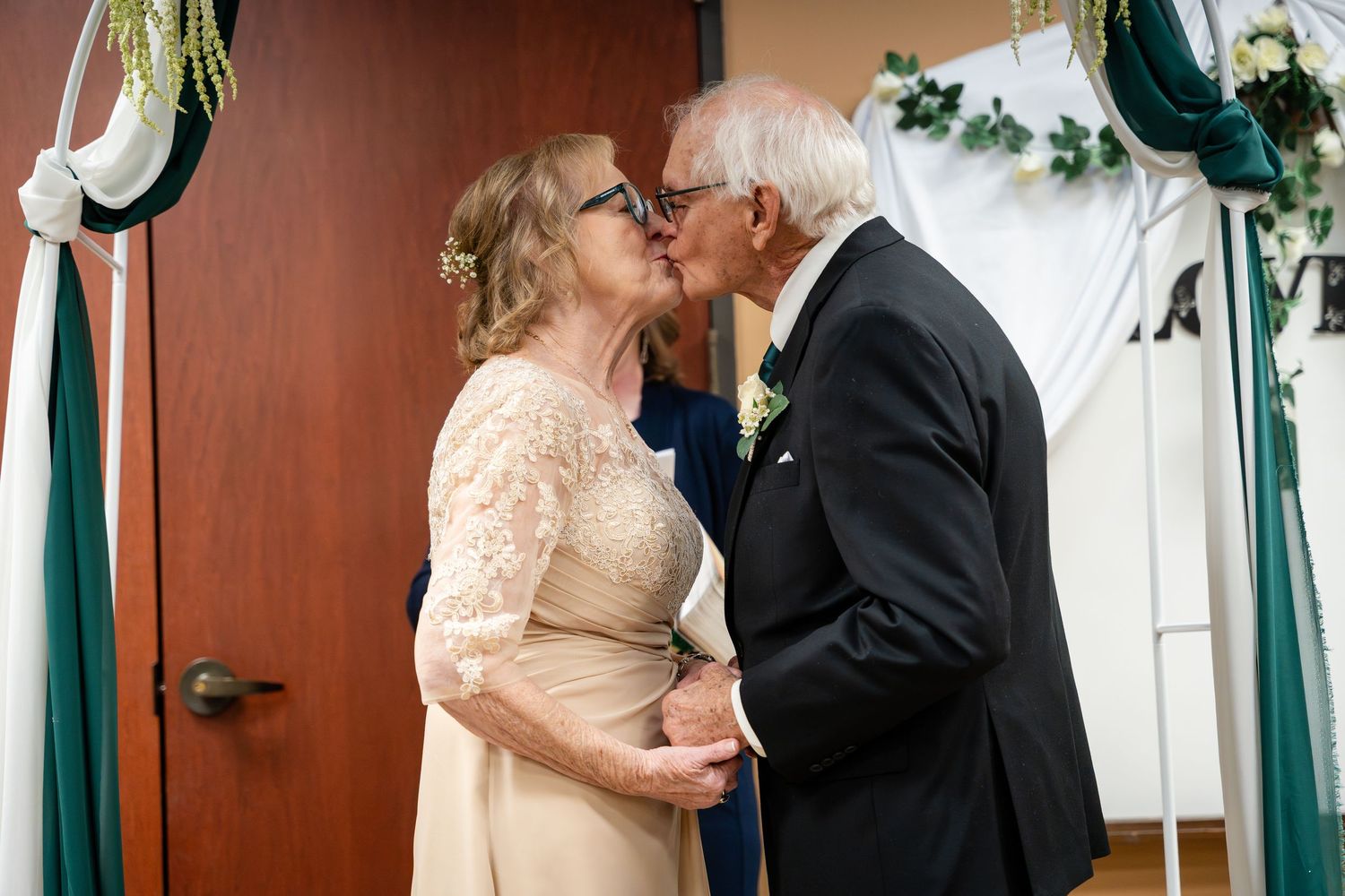 Elderly couple shares a tender kiss at their wedding ceremony with champagne-colored dress and black tuxedo.