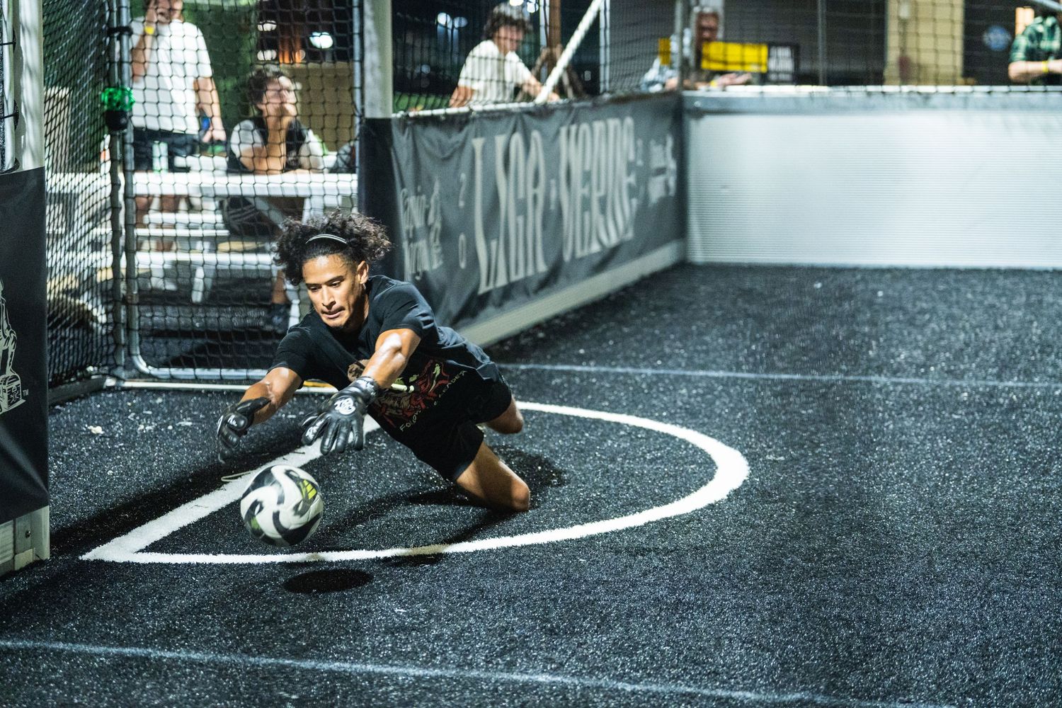 Player dives for the ball during a competitive indoor soccer match on a dark court.