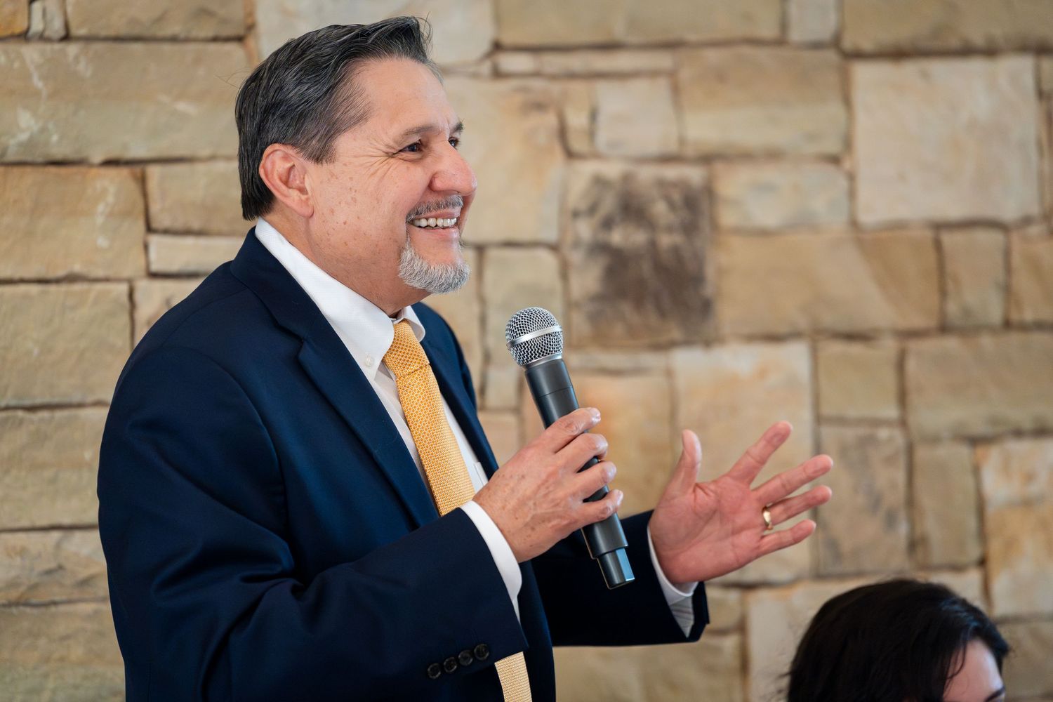Professional speaker in business attire gives animated presentation against stone wall backdrop at event.