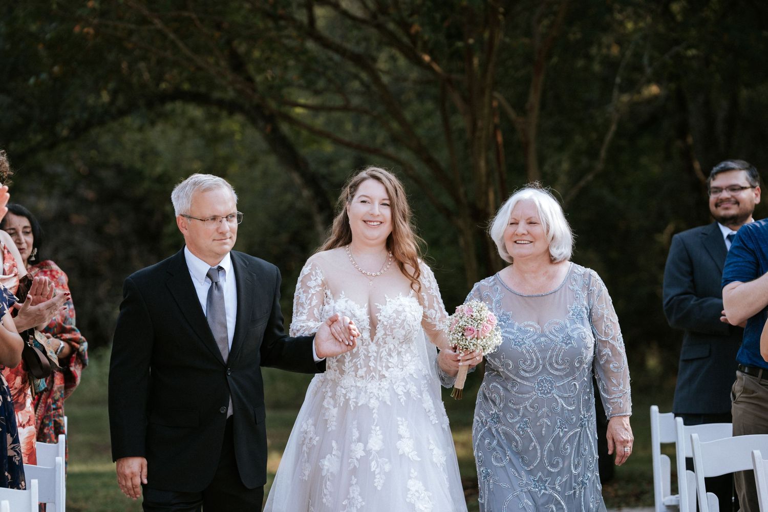 A joyful wedding processional outdoors with the bride wearing a white lace gown walking down the aisle with loved ones.