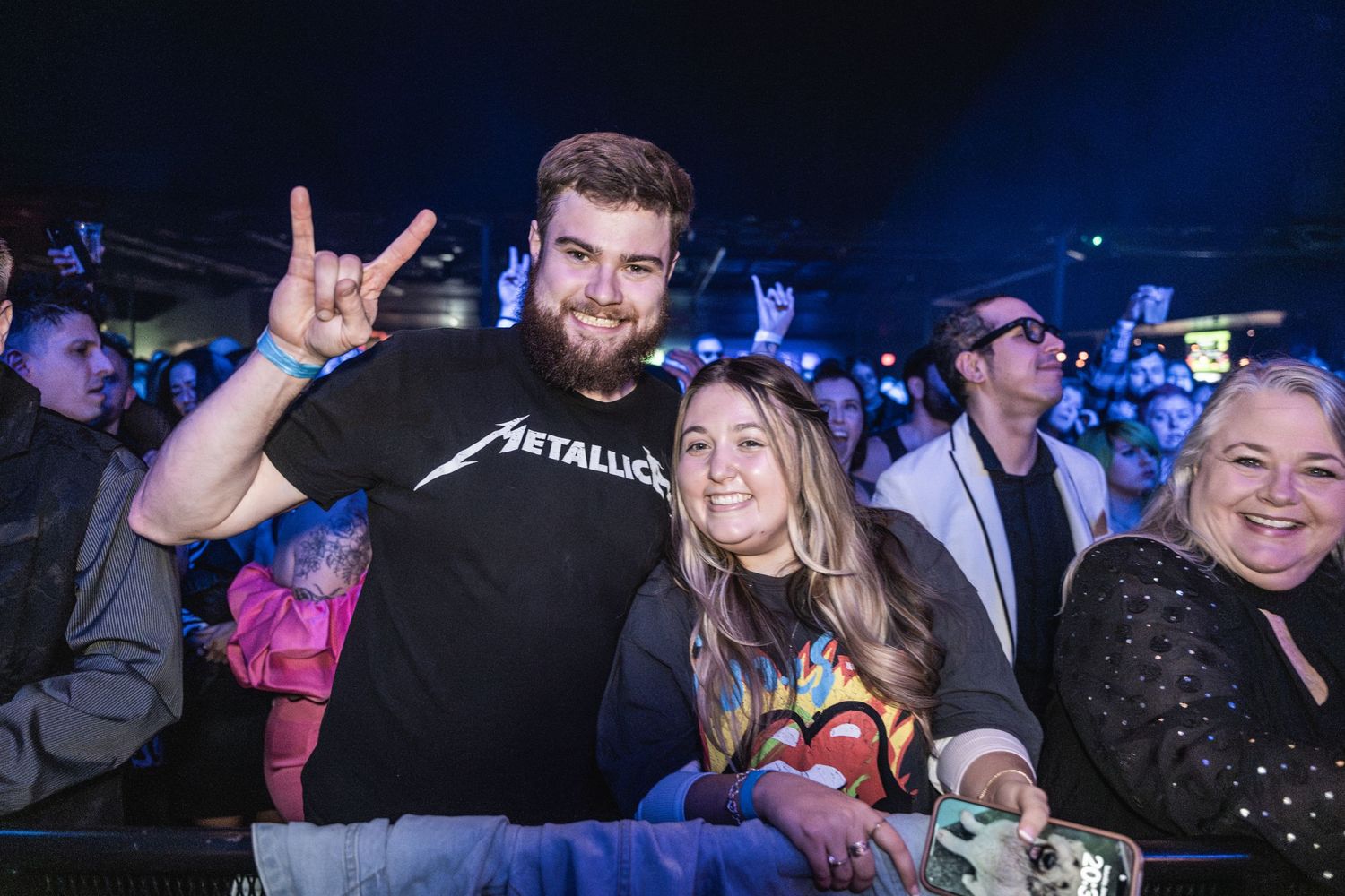 Concert attendees in crowd showing rock hand signs at music show with person wearing Metallica shirt.