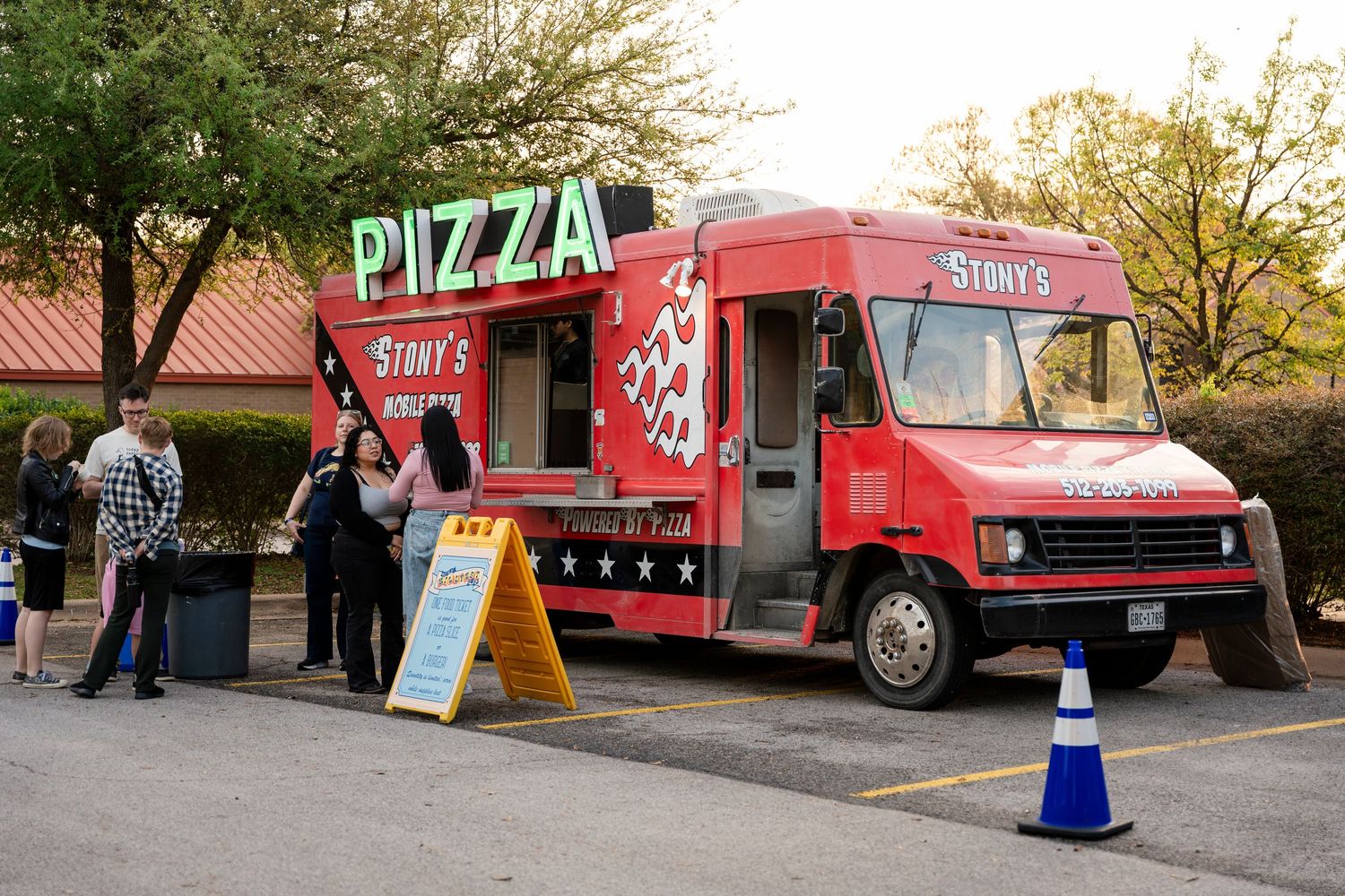 A bright red pizza food truck parked in a lot with customers waiting outside during sunset.