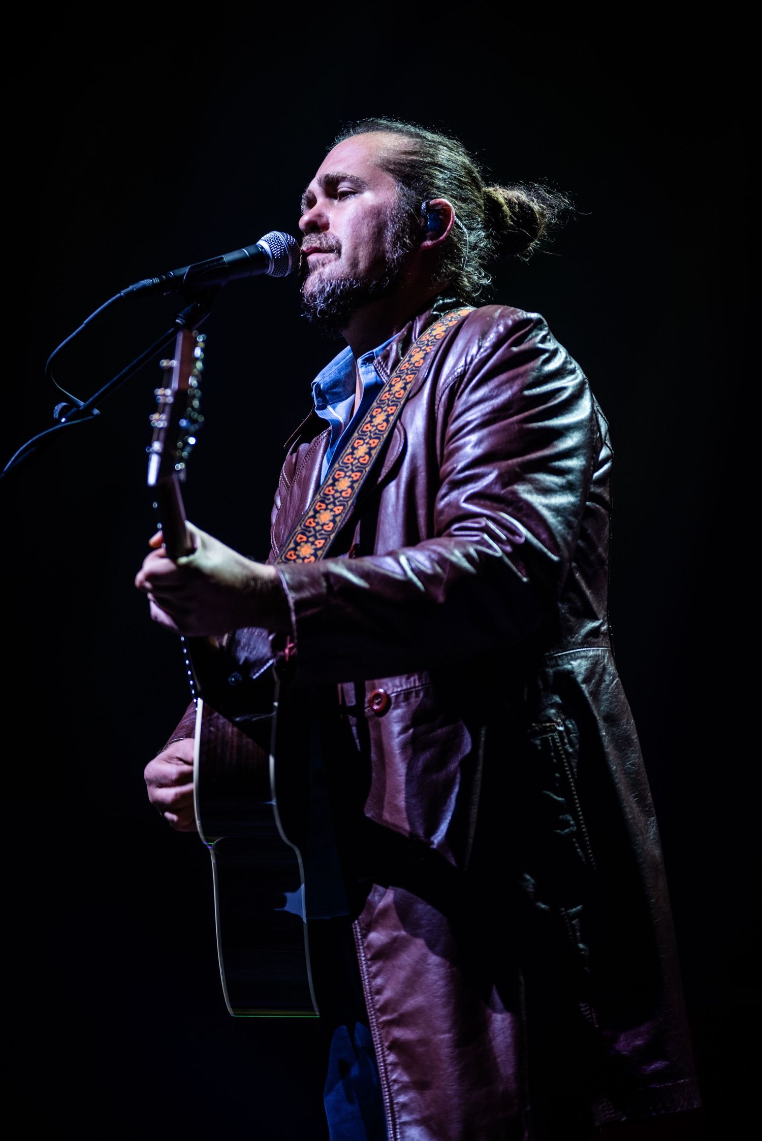 Musician in leather coat performs with acoustic guitar under dramatic stage lighting.