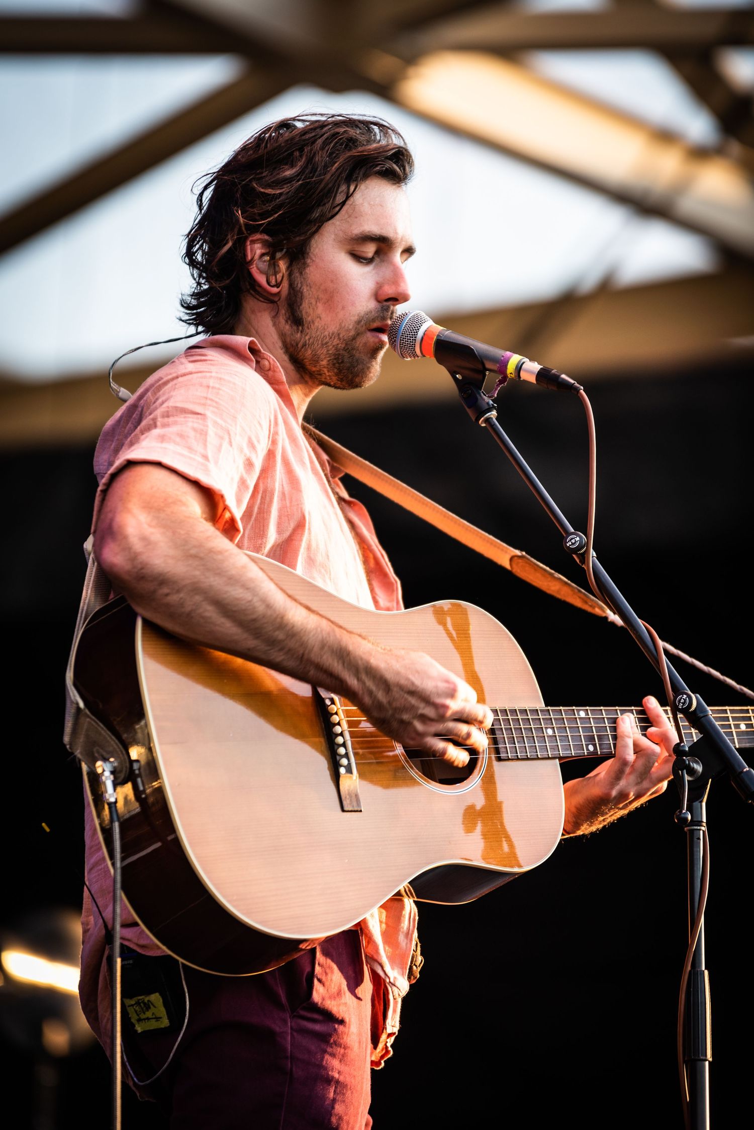 Folk musician strums acoustic guitar while singing into microphone at outdoor venue.