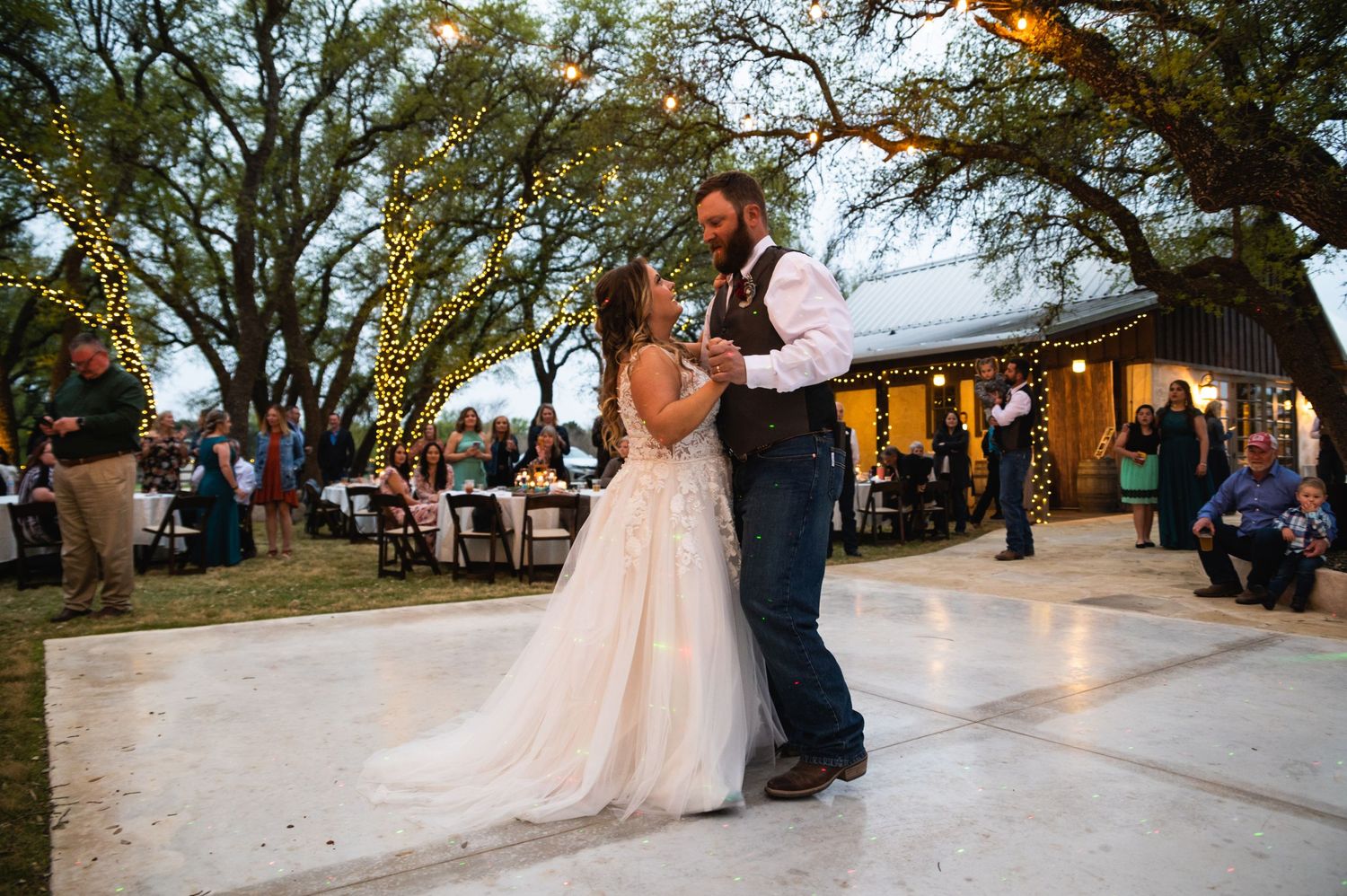 A couple shares their first dance outdoors at a rustic wedding reception with string lights and trees in the background.