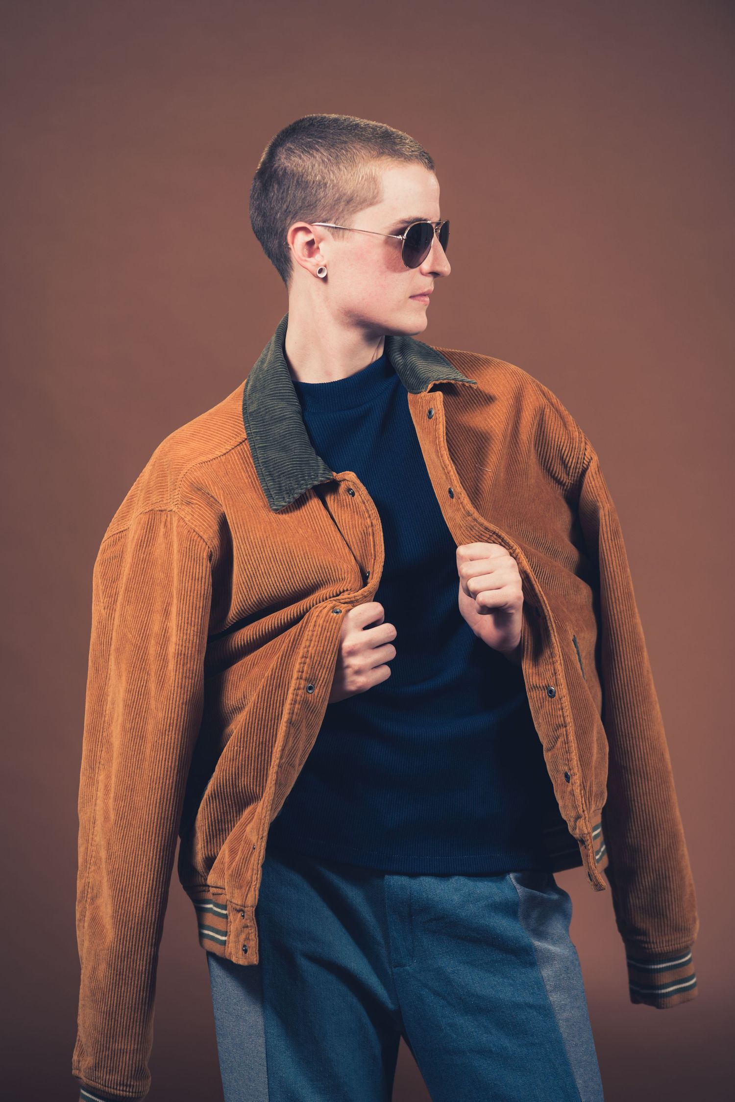Person in navy shirt and corduroy jacket wearing sunglasses against brown background.