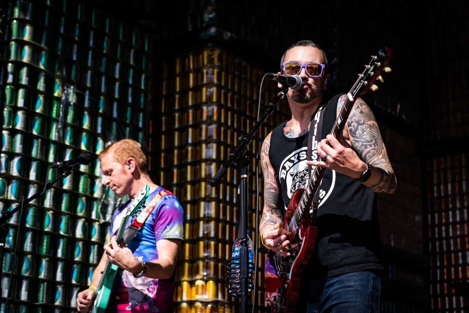 Two musicians perform on stage with guitars against a dark backdrop with green lighting.