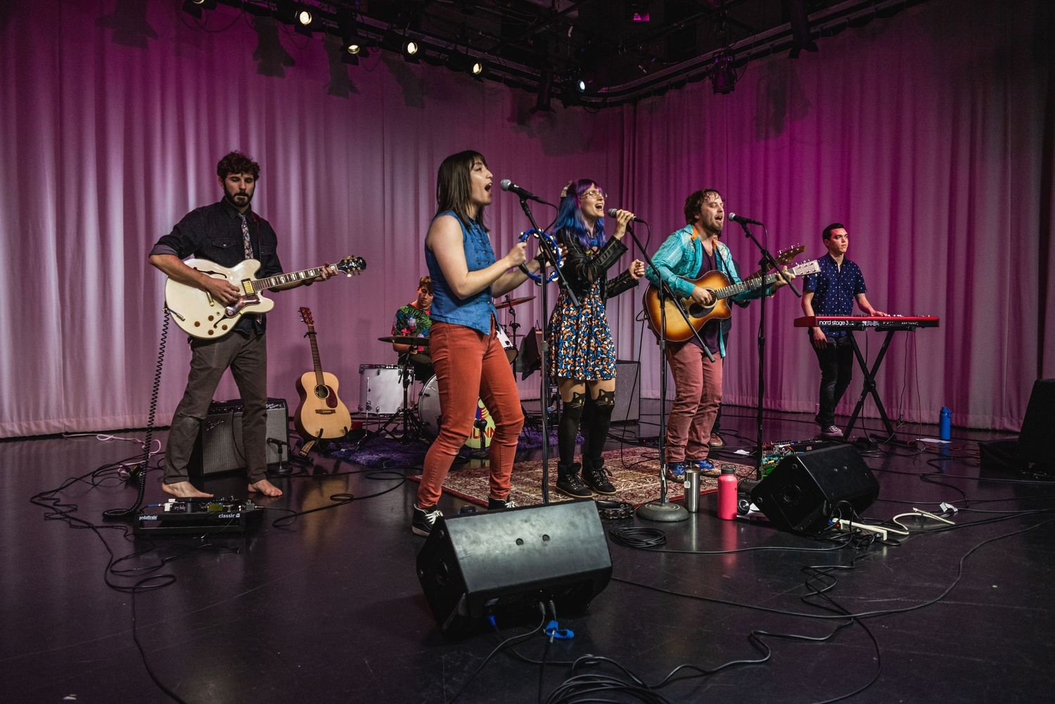 Full band performs on dark stage with multiple instruments and professional lighting.