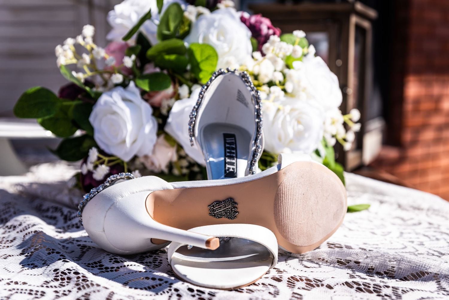 Bridal shoes and floral arrangement displayed on white lace tablecloth in natural light.