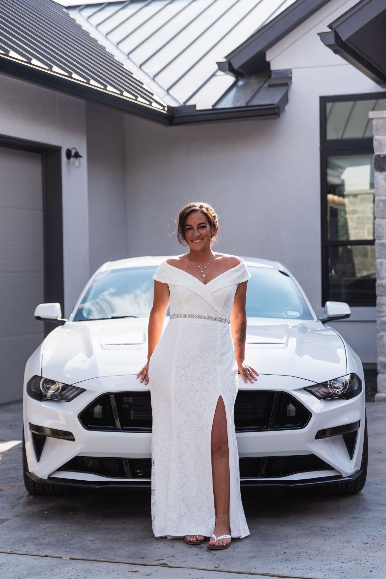 Elegant white wedding dress with slit poses in front of silver Ford Mustang sports car.