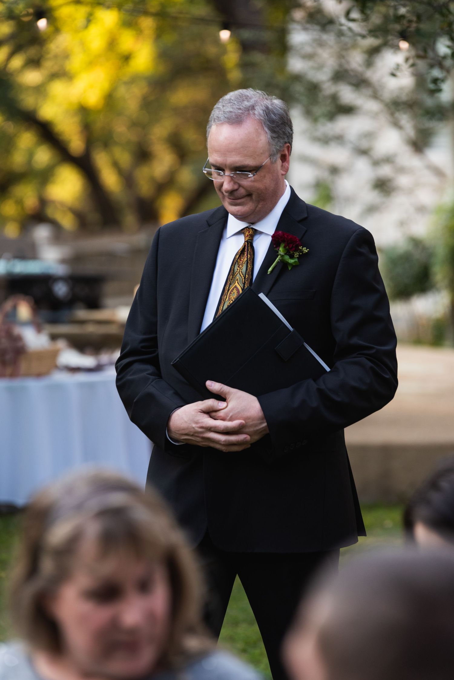 An officiant in a black suit holds papers during an outdoor wedding ceremony.