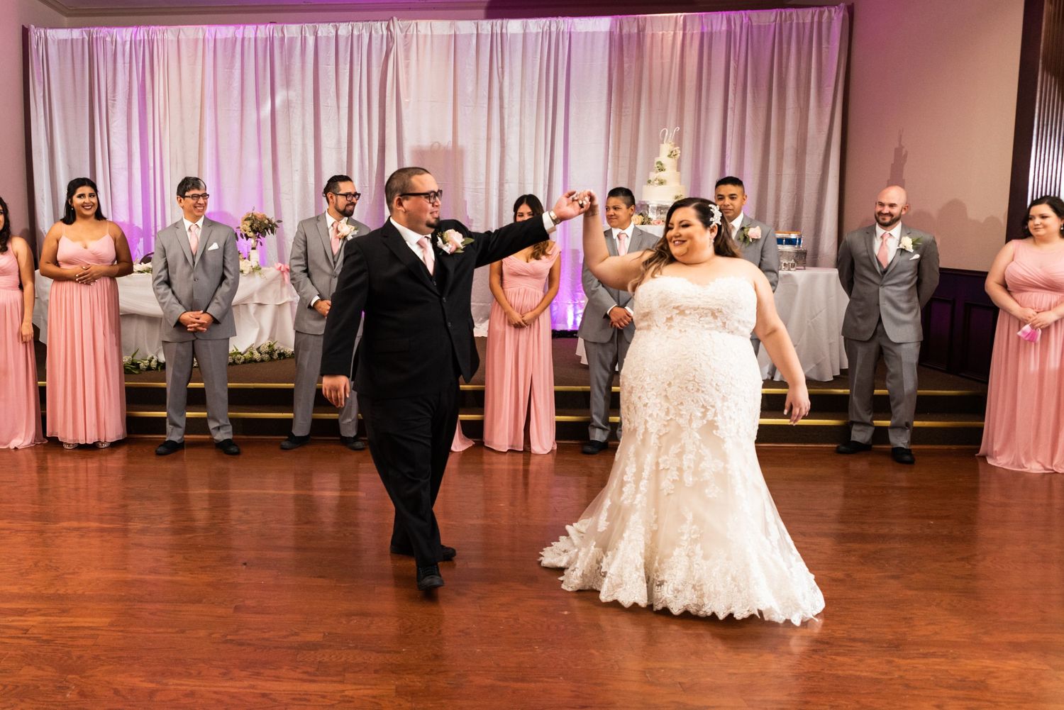 Wedding couple dancing in a reception hall with bridesmaids in pink dresses and groomsmen in background.