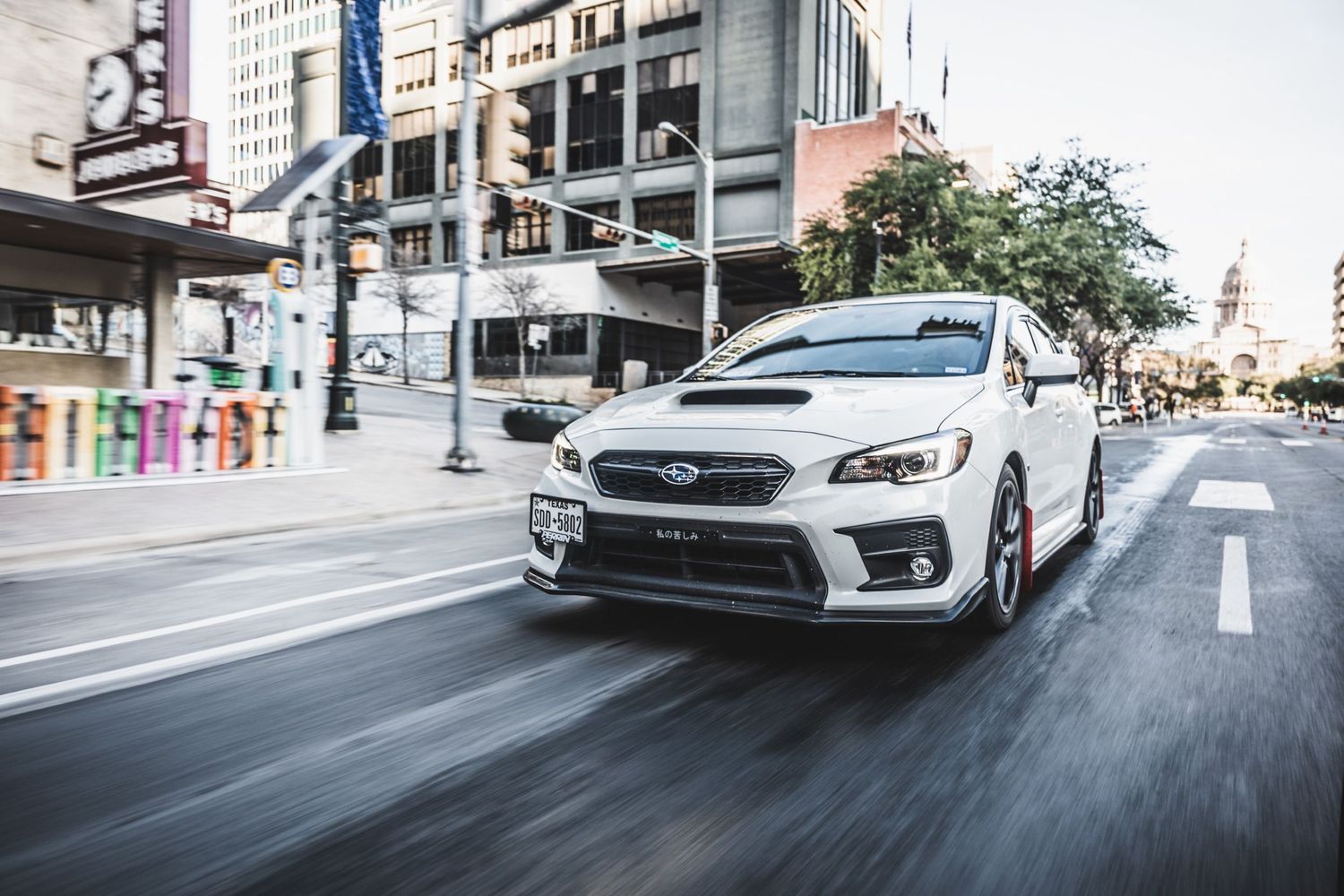 White Subaru WRX STI sports car driving through downtown streets with motion blur effect.