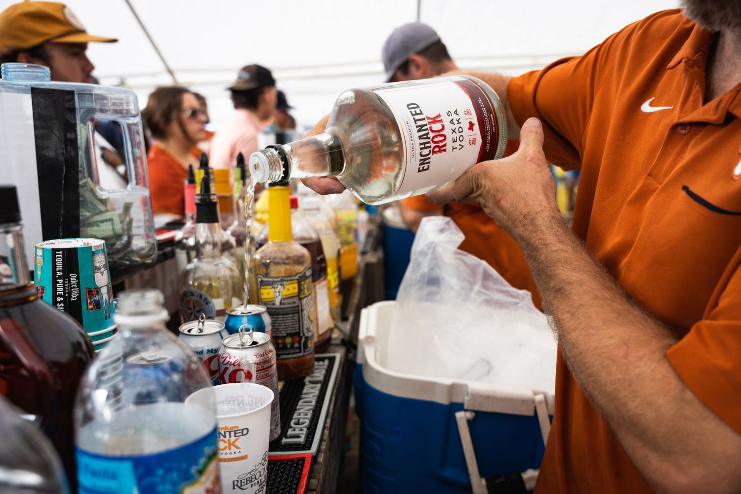 Someone pours liquid from a bottle into a container at an outdoor bar station.