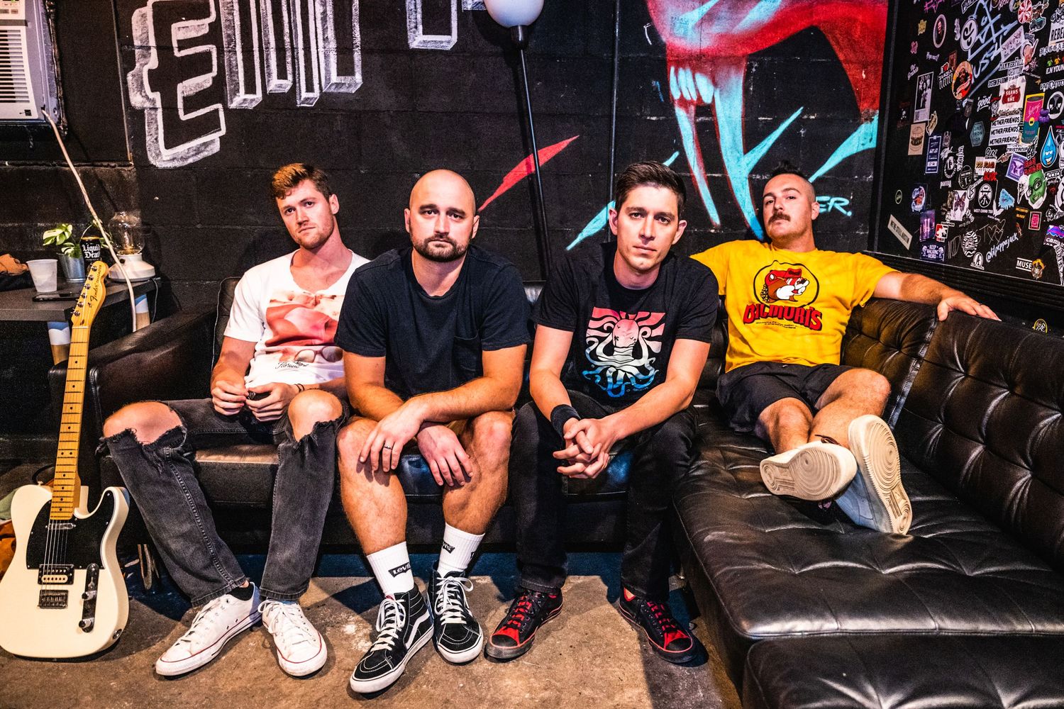 Rock band members relaxing on leather couch in music studio with instruments.