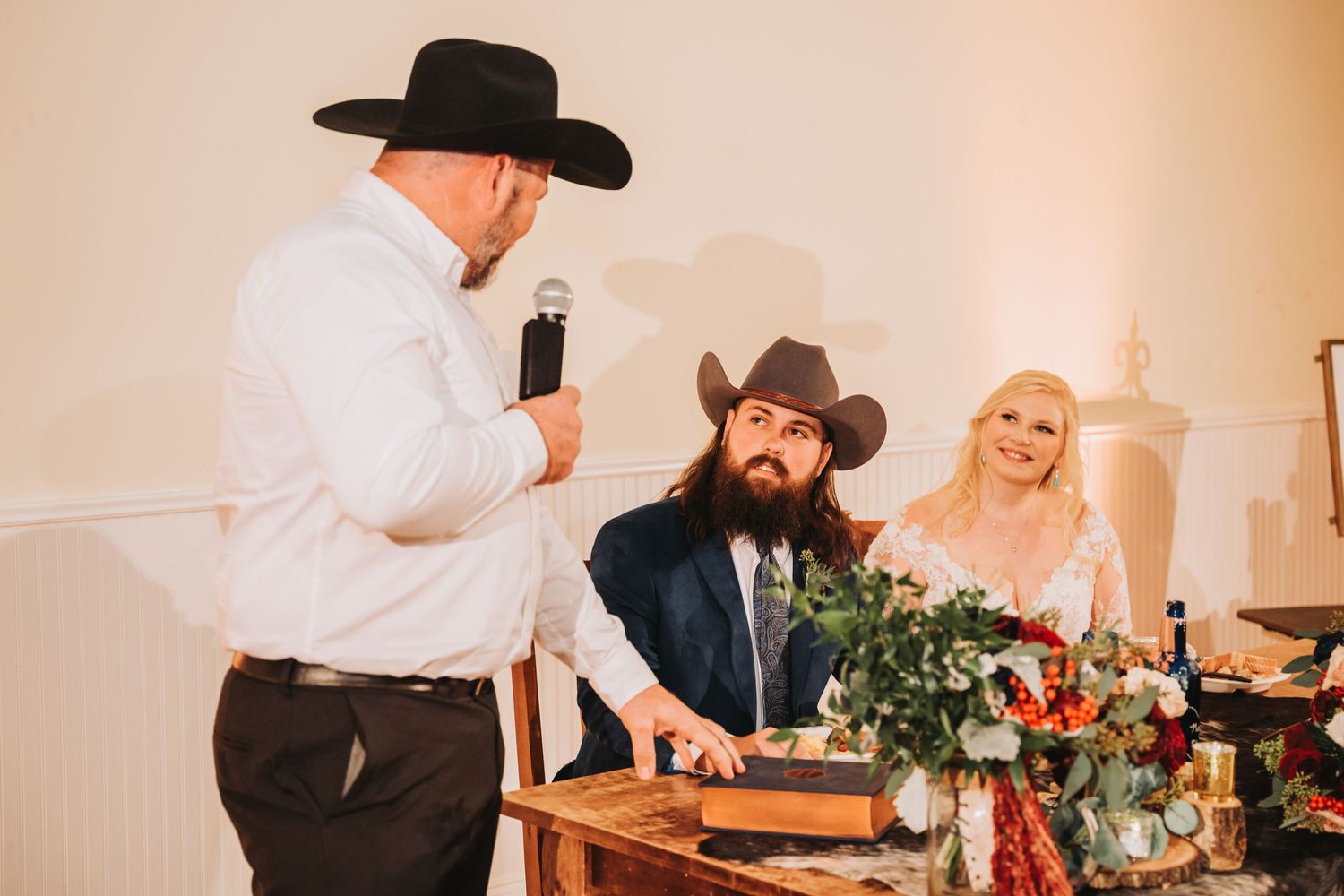 A wedding reception scene with guests gathered around decorated tables during speeches.