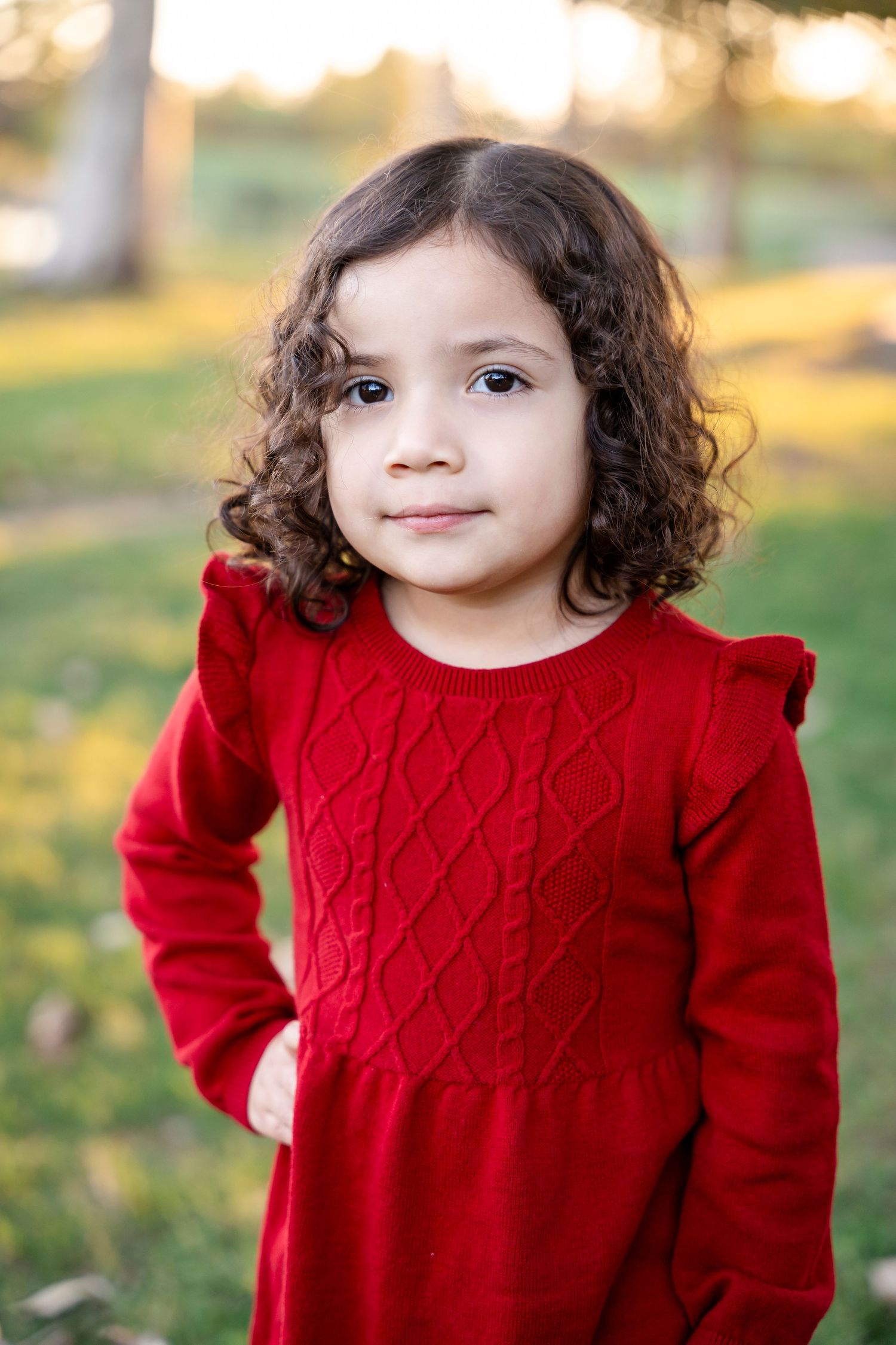 Young person in red knit dress with curly hair outdoors at sunset.