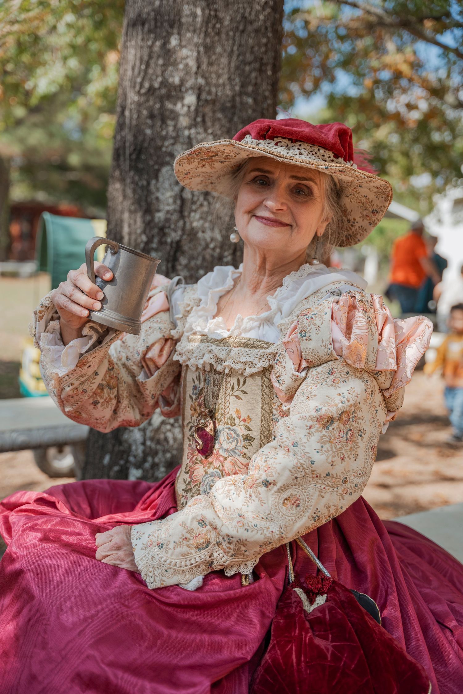 A person in an ornate Victorian dress and hat holds a cup while sitting outdoors.