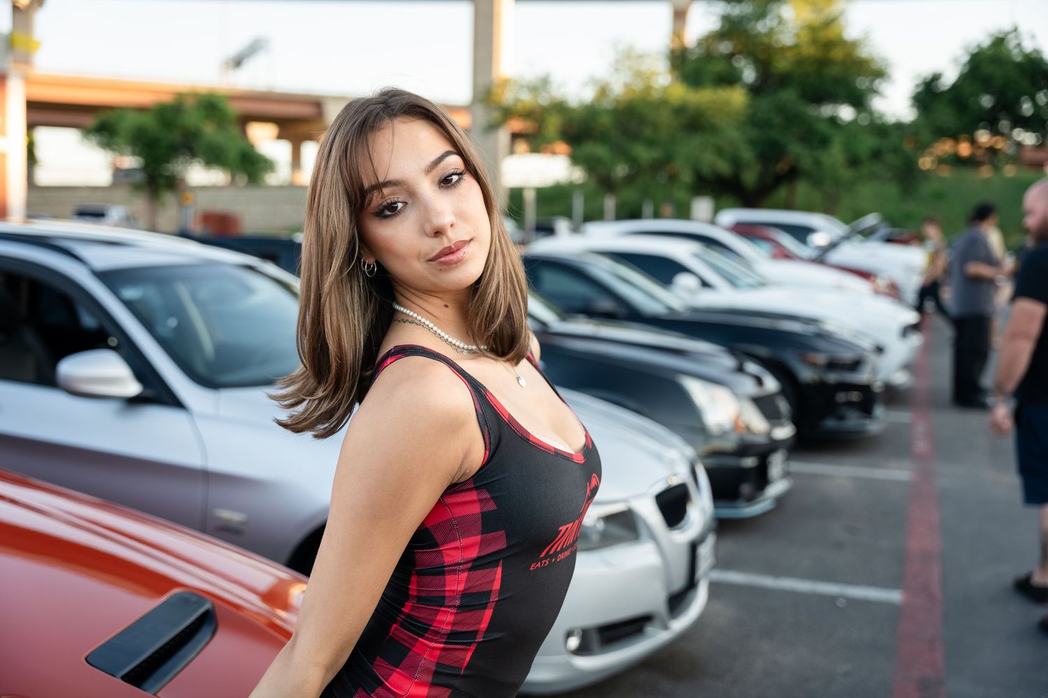 Model in black and red outfit poses against cars at outdoor auto show.