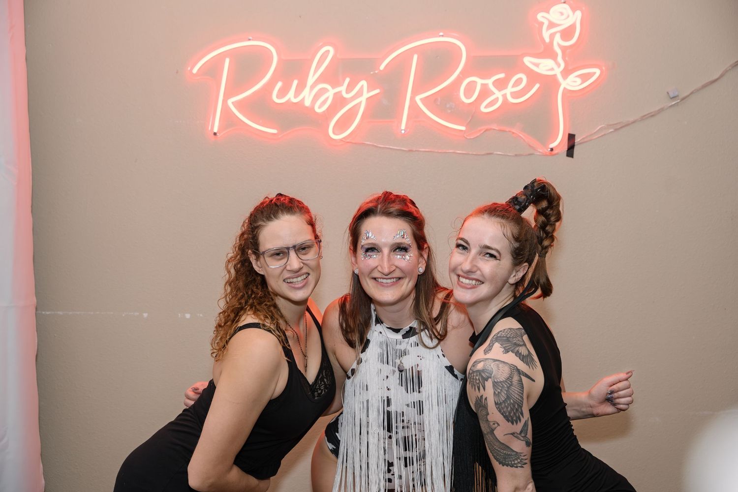 Three people in black outfits posing under pink neon Ruby Rose sign.