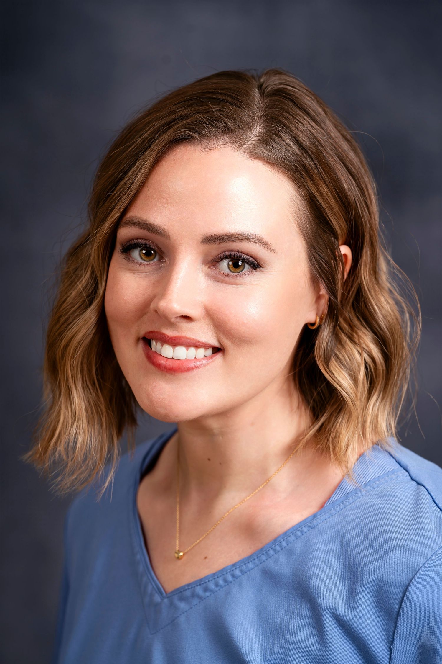 Professional headshot of woman in blue scrubs against dark background.