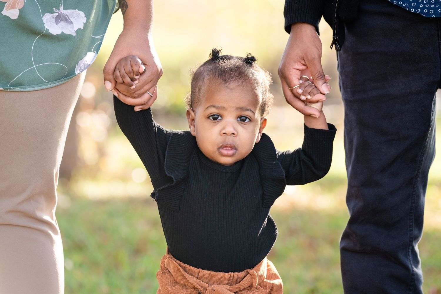 A small child in black top and brown pants learning to walk while holding hands.