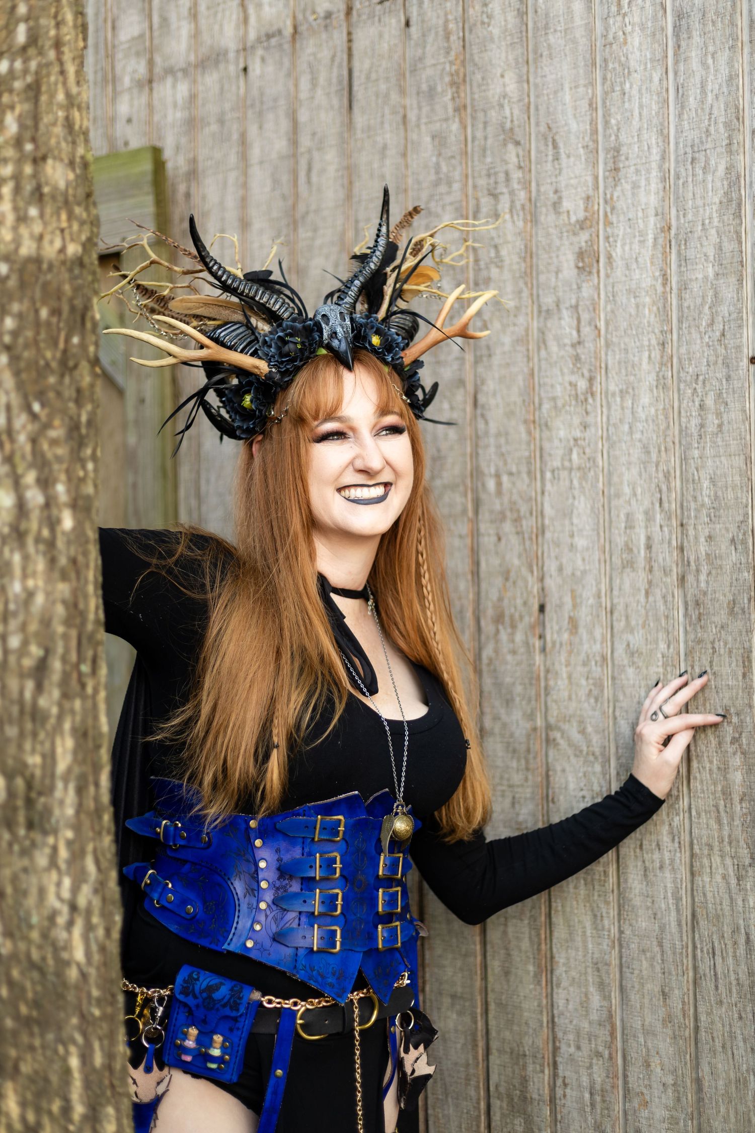 Person in a blue corset and feathered headdress poses playfully against a rustic wooden wall.