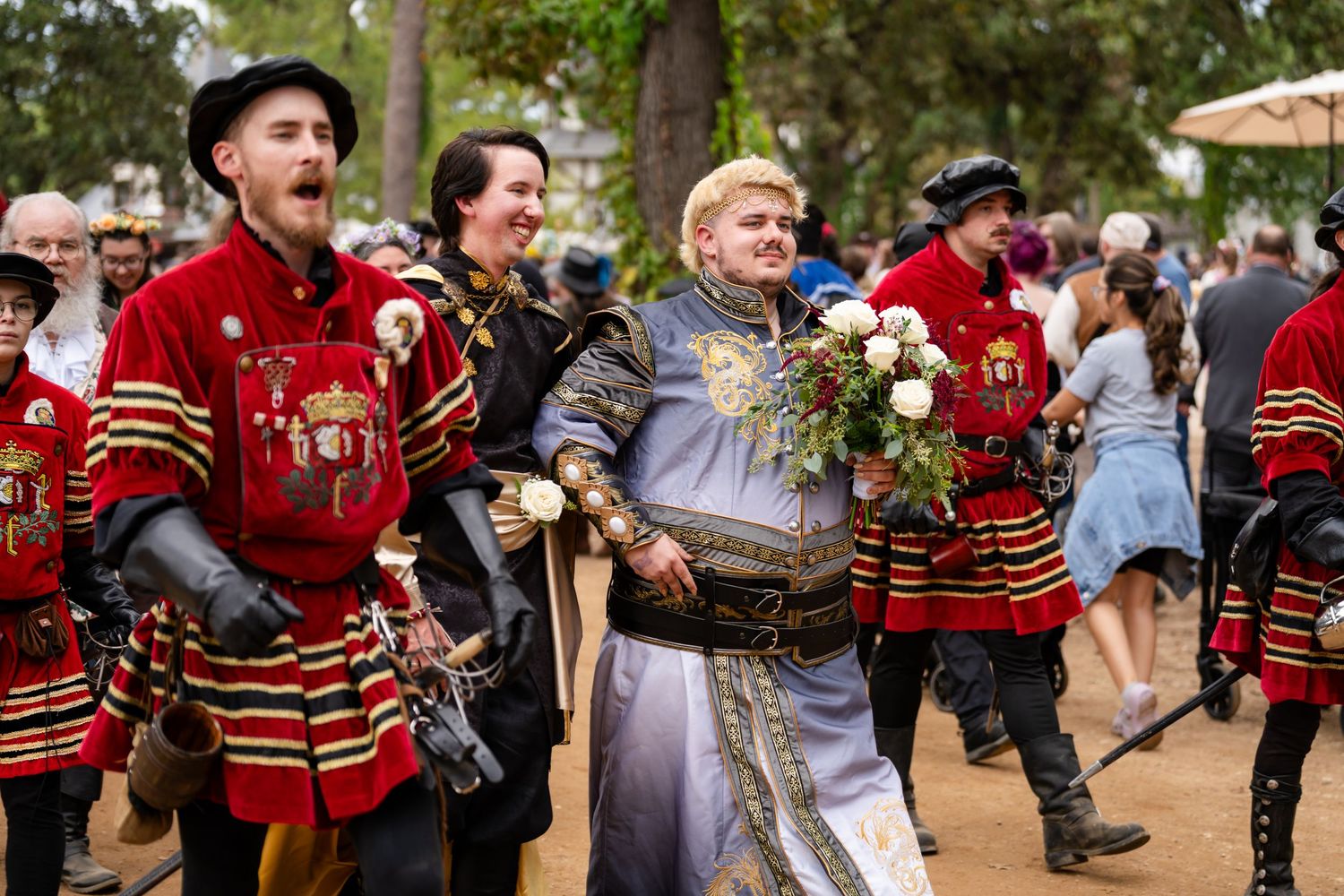 Performers in ornate red and black period costumes celebrate at a Renaissance festival with joyful expressions.
