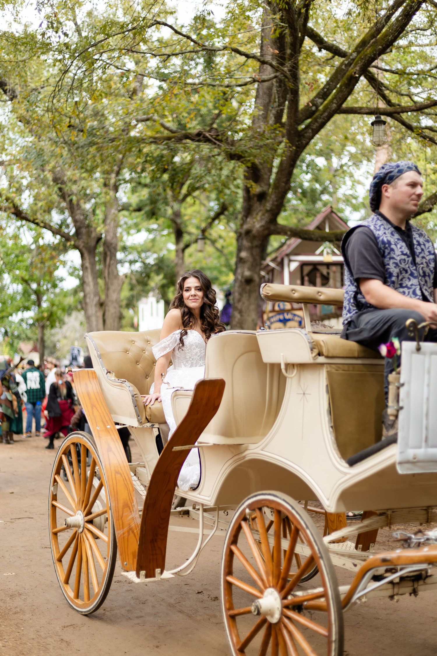 A white horse-drawn carriage with wooden wheels carries passengers through a tree-lined path at a park.