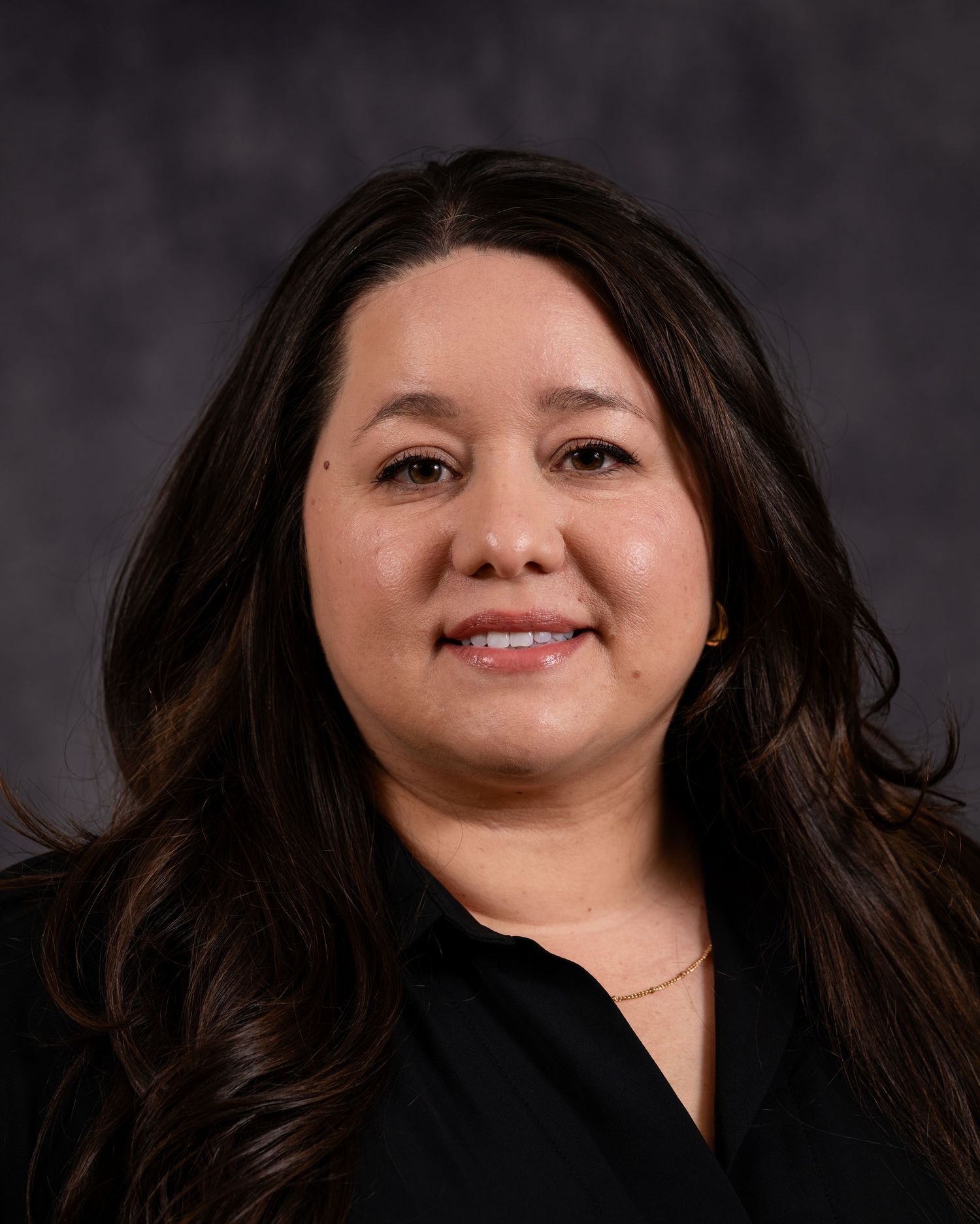 Professional headshot of a person with long dark hair wearing a black top against a dark gray background.