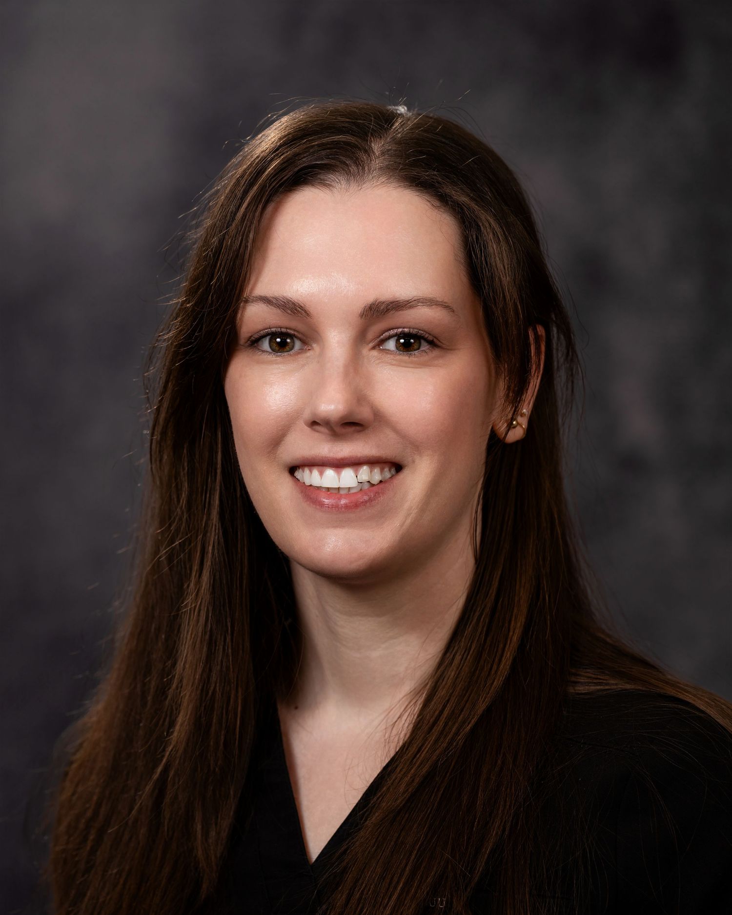 Professional headshot portrait with dark background showing a warm smile and long brown hair.