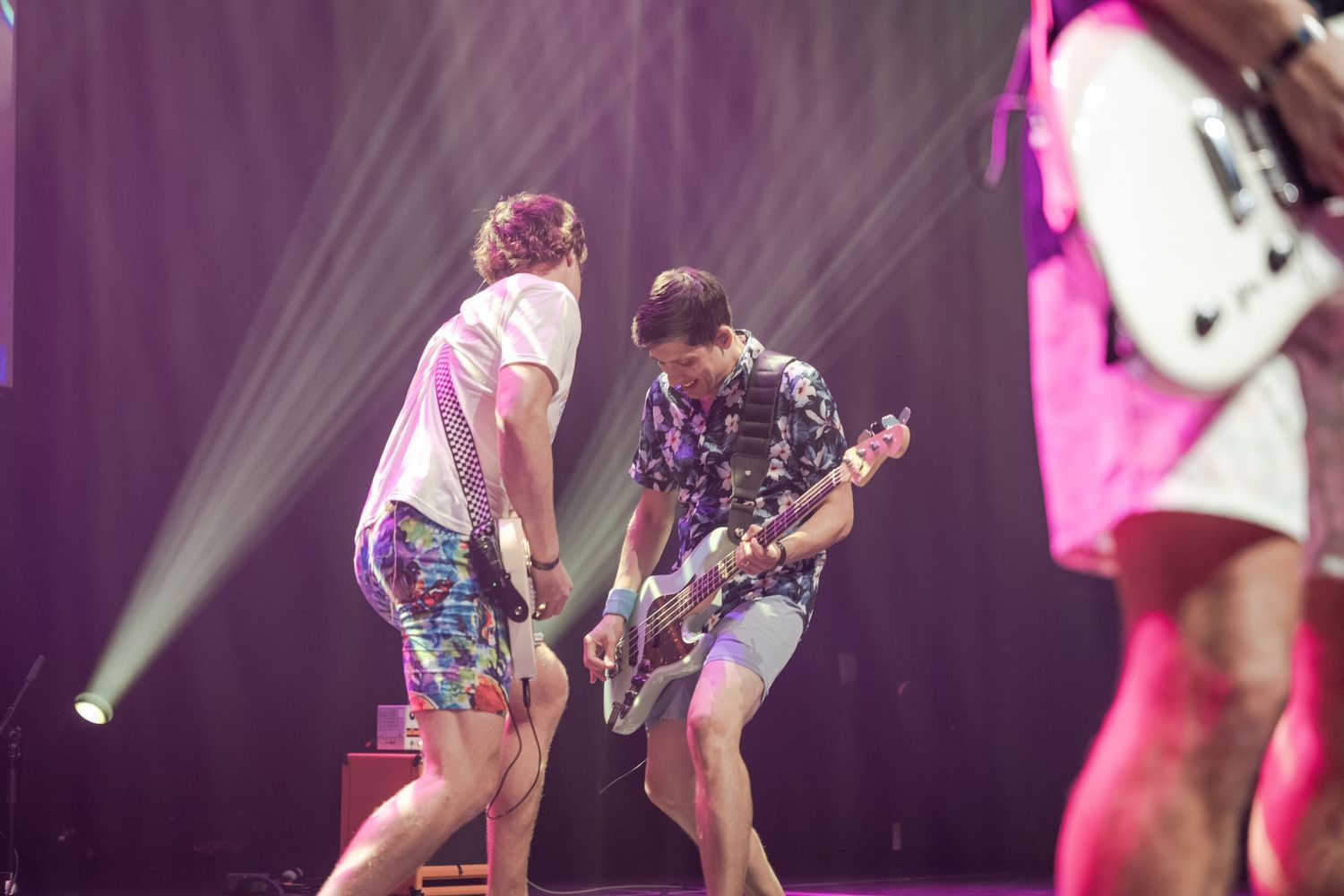Two musicians perform on stage under bright lights, playing guitar in colorful summer outfits at a live concert.