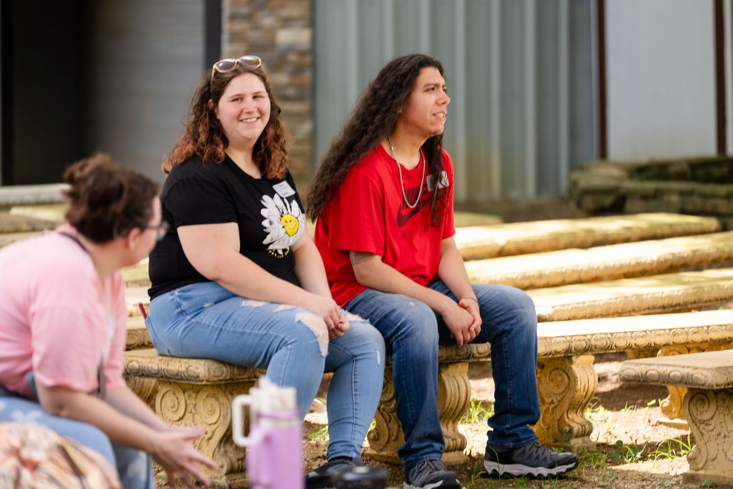 Two students sit on a decorative stone bench outdoors on a college campus on a sunny day.