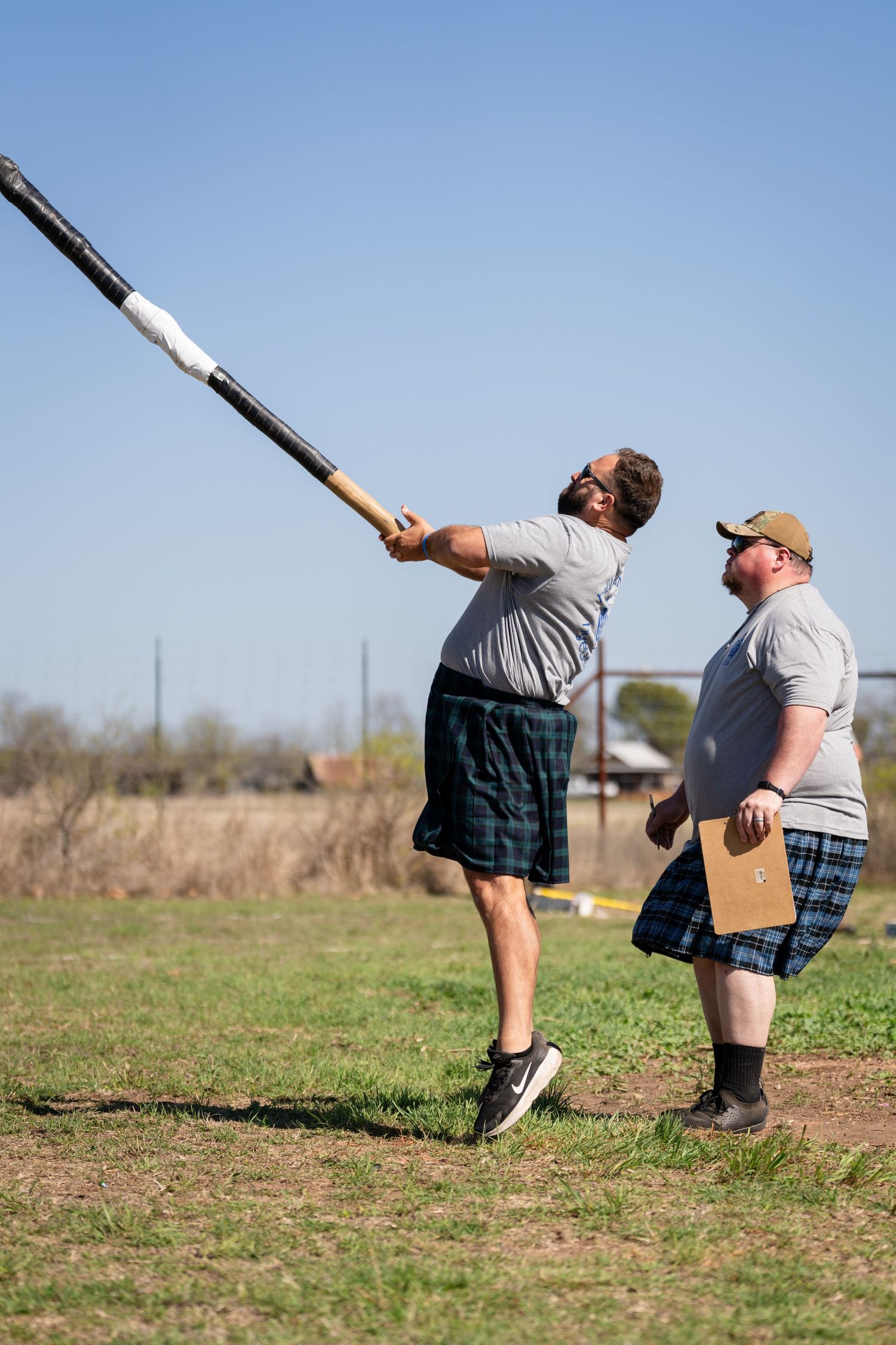 Two men in plaid shorts compete in a caber toss at an outdoor Highland Games event on a sunny day.
