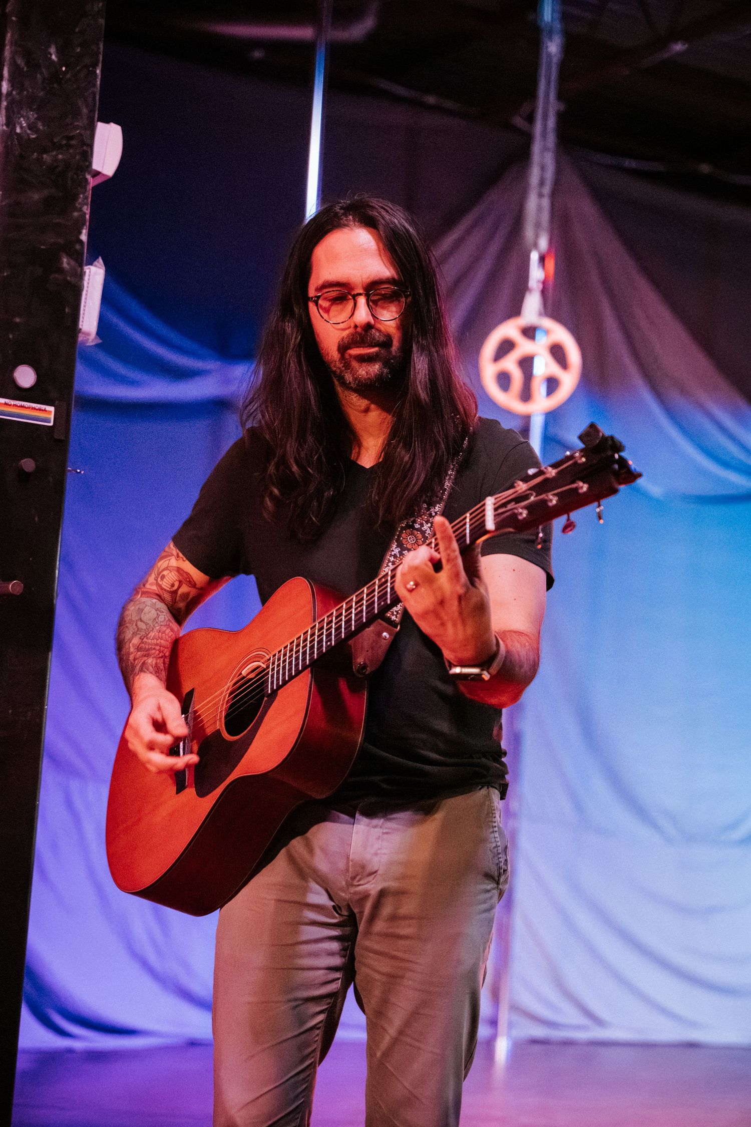 Long-haired guitarist with tattoos performs on acoustic guitar under blue stage lighting at an indoor venue.