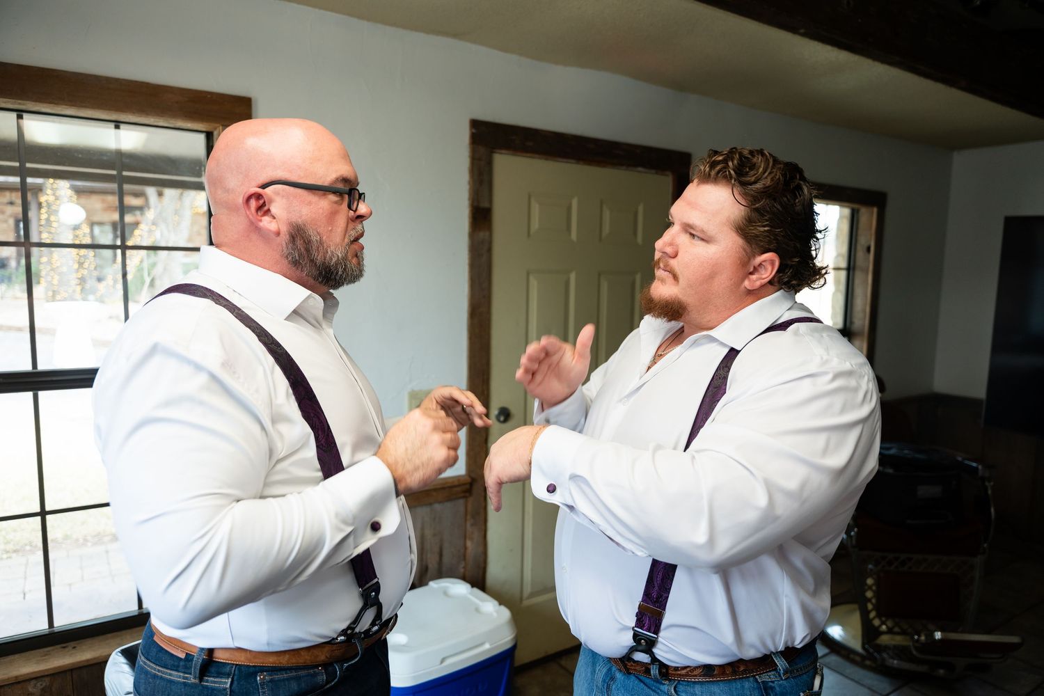 Two groomsmen in white shirts and suspenders help each other with cufflinks before a wedding ceremony.