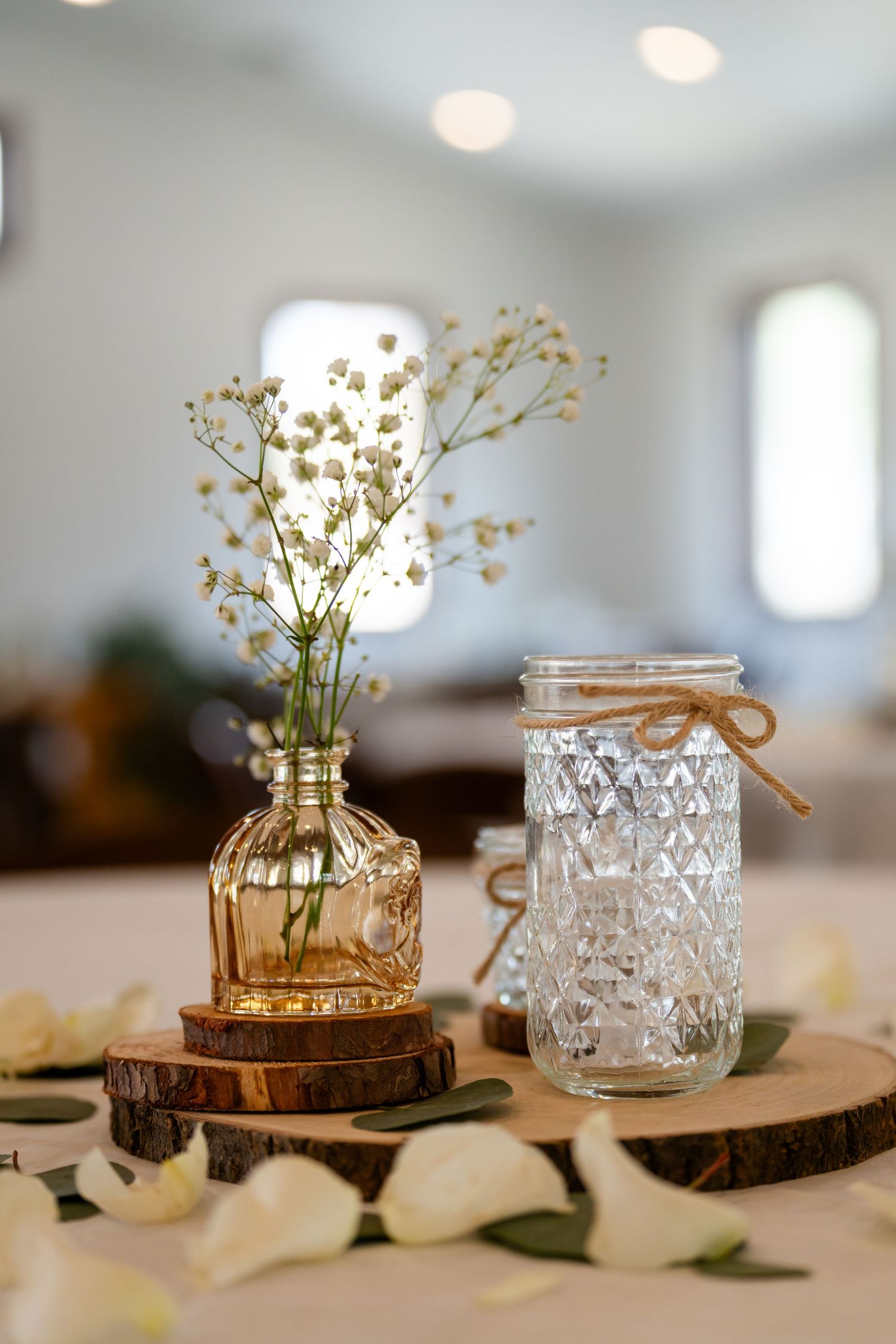 Rustic wedding centerpiece with baby's breath in amber bottle and mason jar on wood slice with white petals.