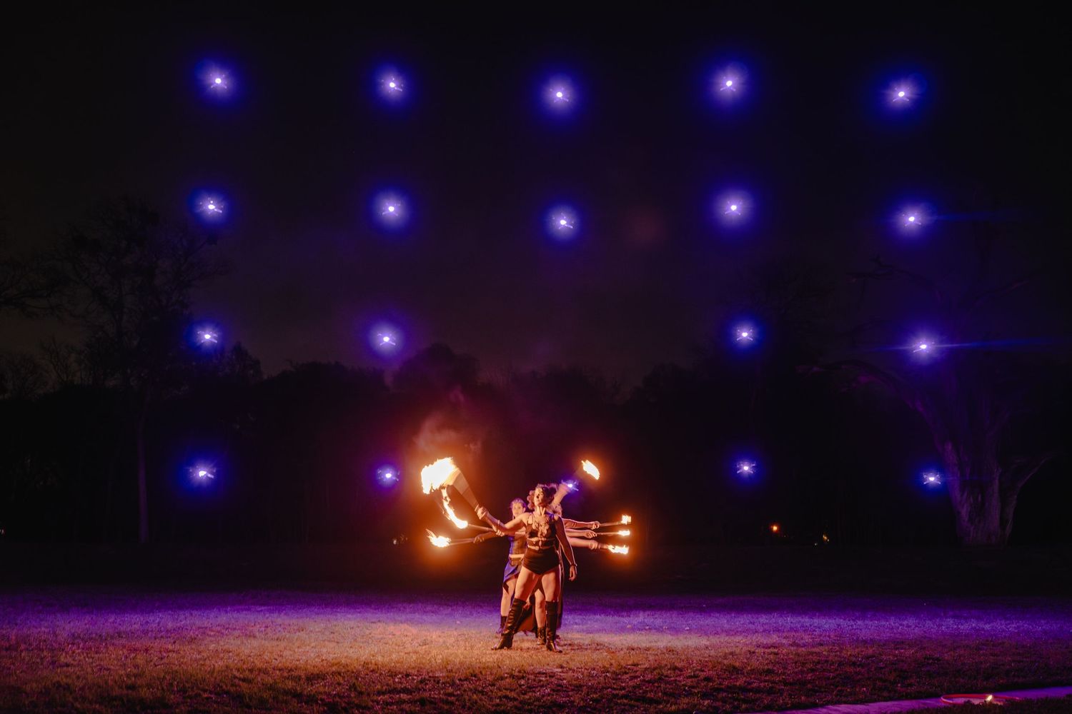 Fire dancer performing at night with glowing blue lights backdrop, wielding flaming torches in an outdoor show.