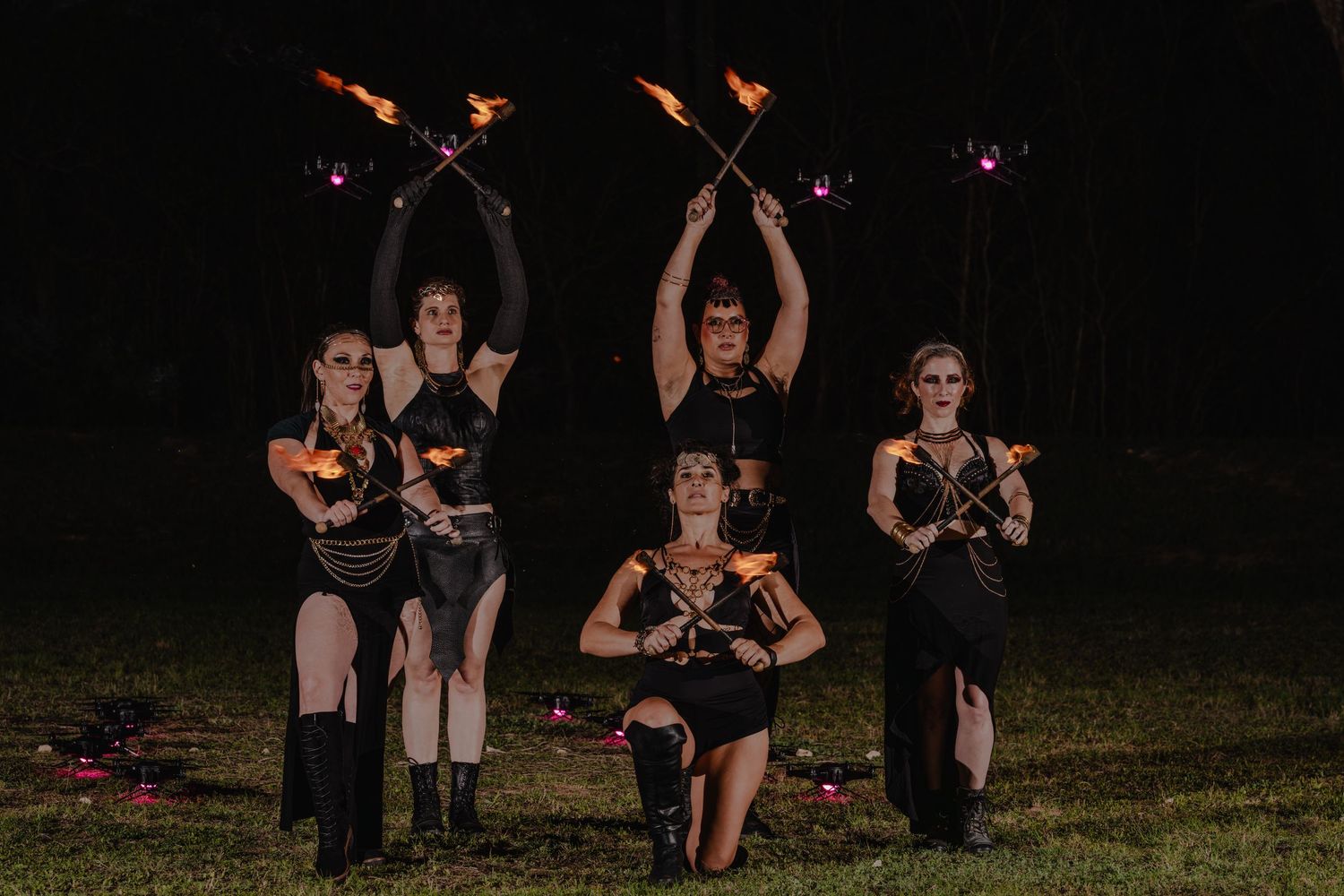 Five performers in black costumes hold flaming props during a dramatic nighttime fire dance show outdoors.