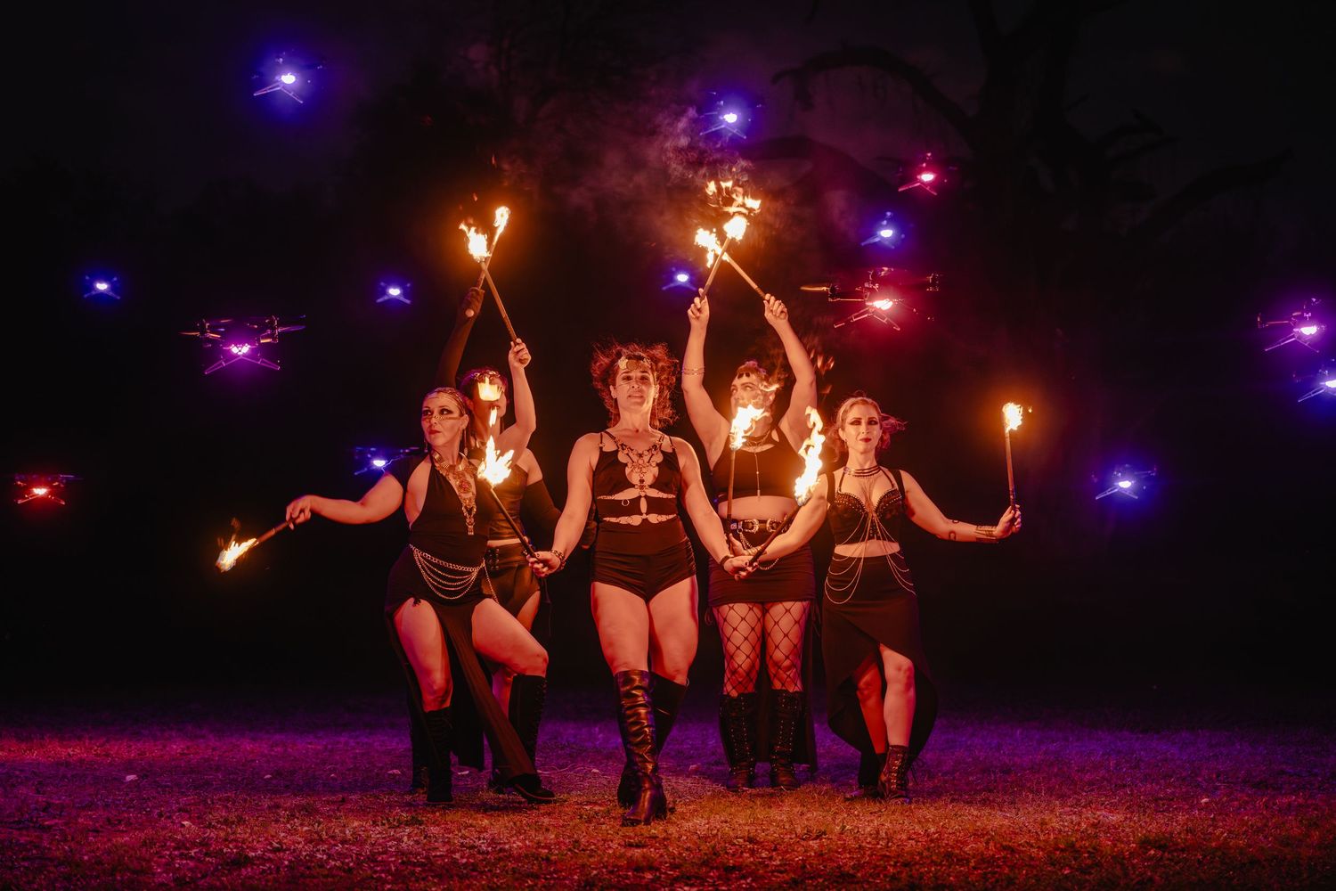 Five female fire performers pose dramatically at night holding torches and sparklers with colorful lights glowing behind them.