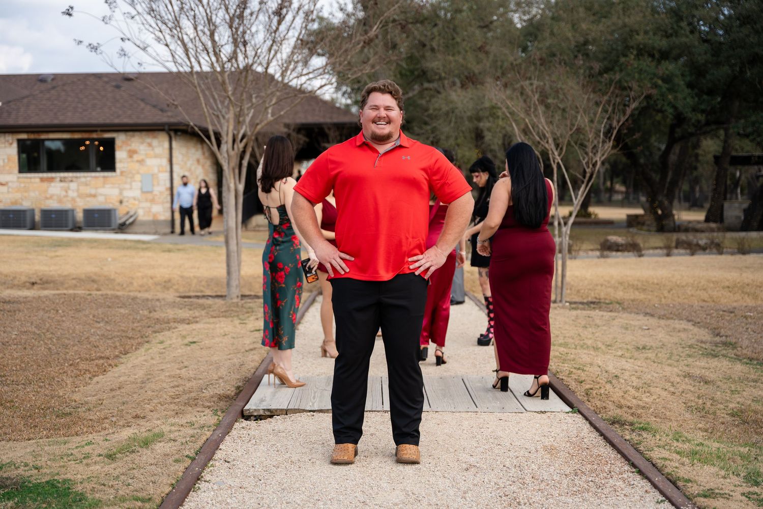 Man in red polo shirt stands confidently outdoors at a rustic venue with guests in formal attire in background.