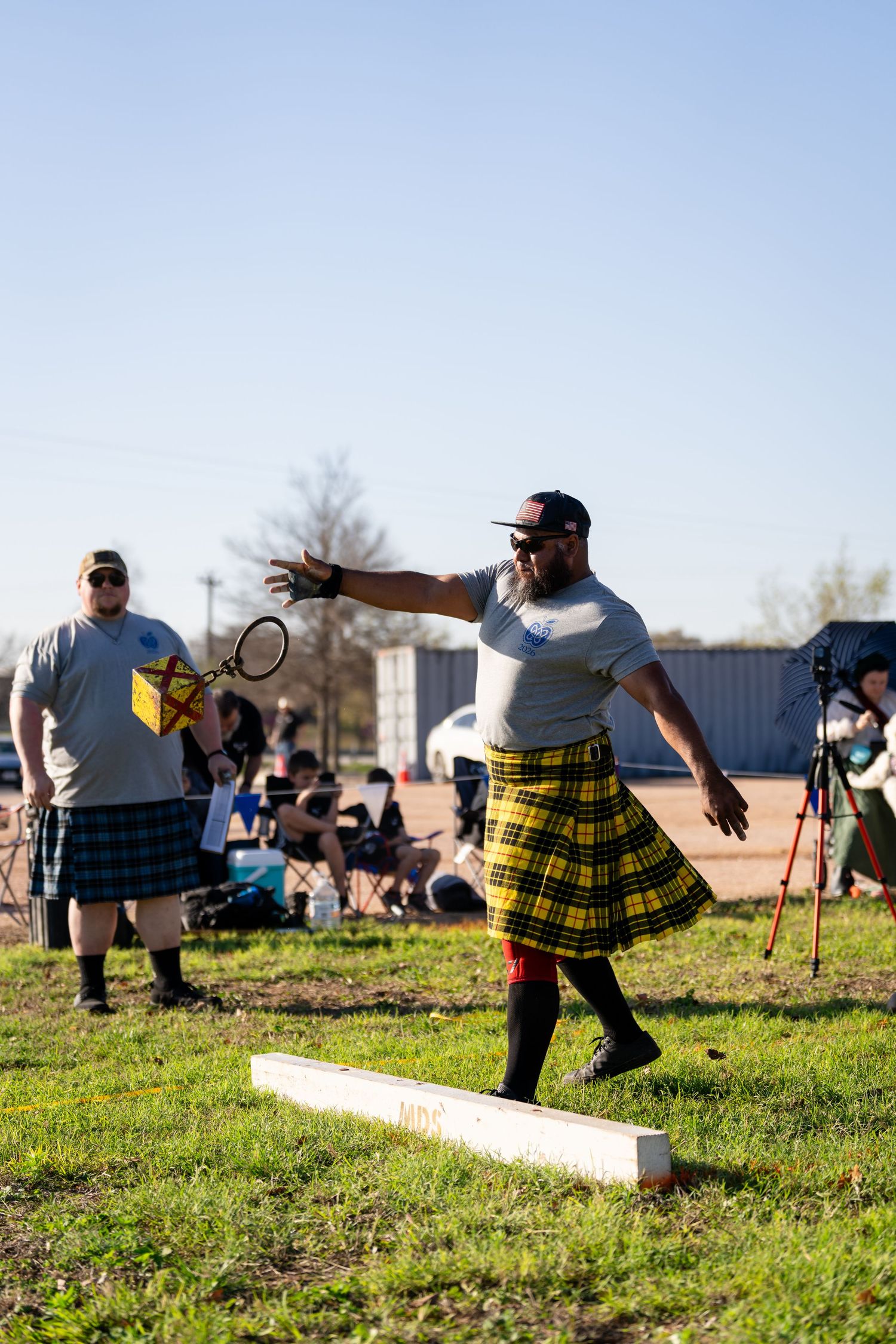 Man in yellow tartan kilt throwing at Scottish Highland Games competition on green field.