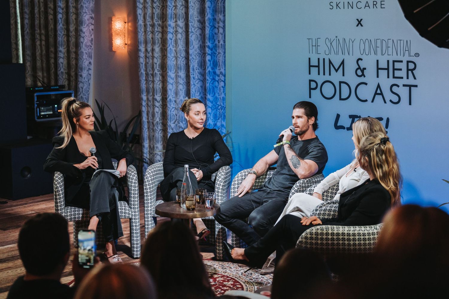 People seated in chairs on stage during a podcast recording session with studio lighting and microphones.