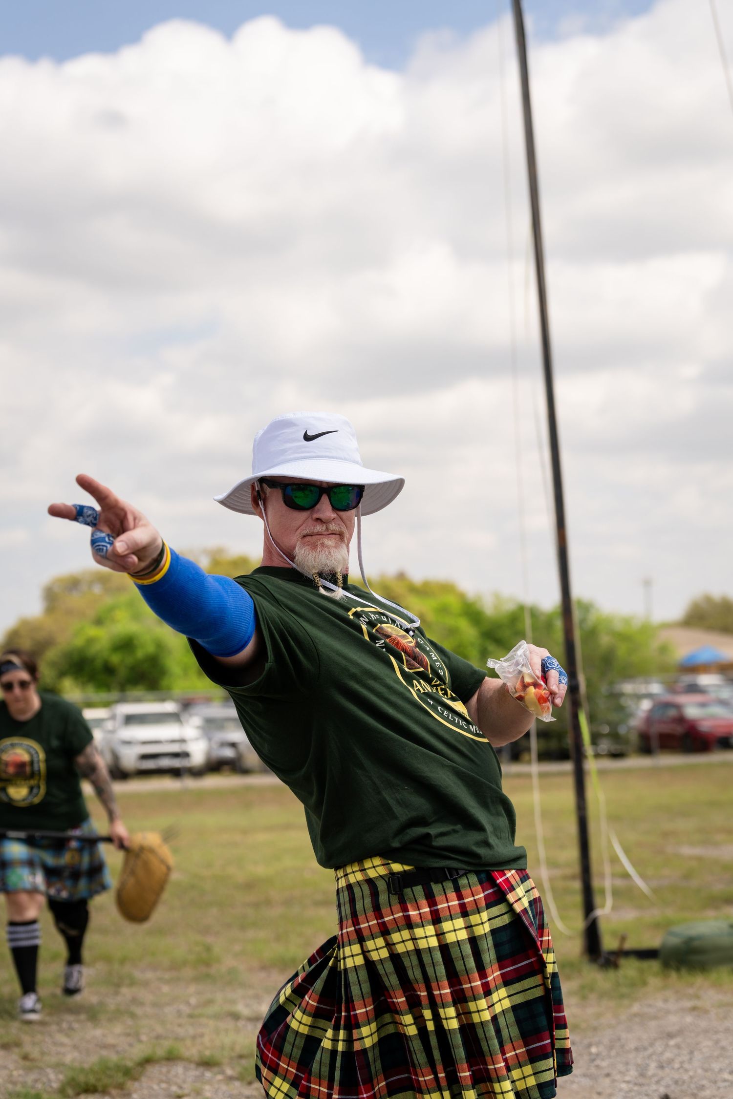 Man in tartan kilt and white Nike hat throwing object at Scottish Highland Games outdoor competition.