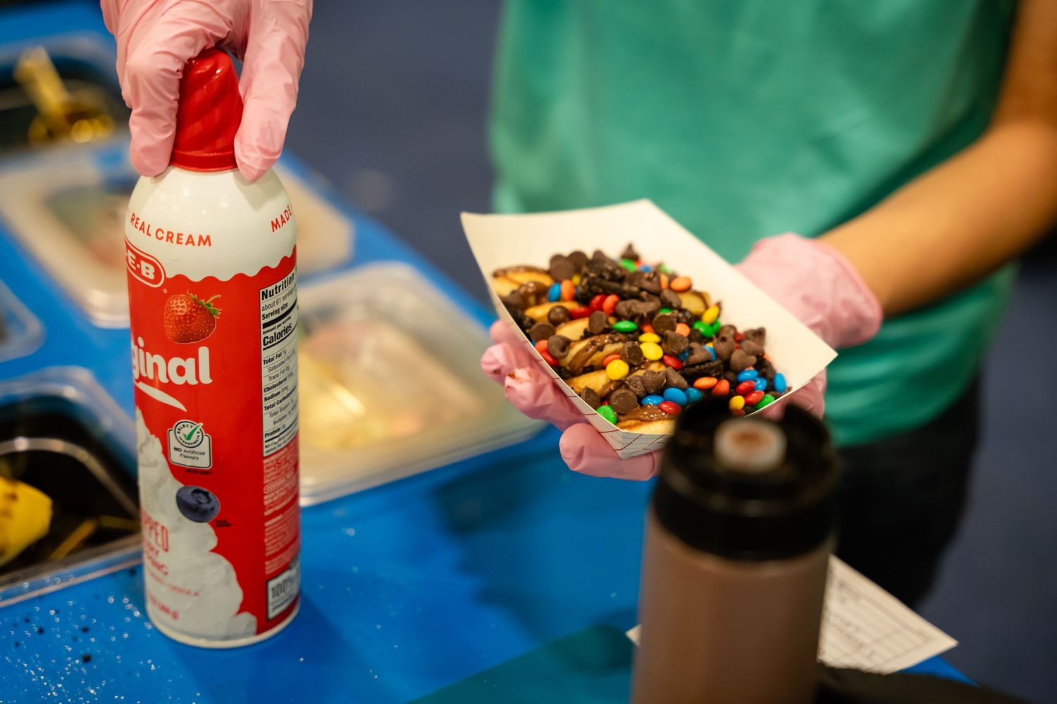 Person holding dessert topped with M&Ms and chocolate drizzle, with whipped cream can on blue counter.