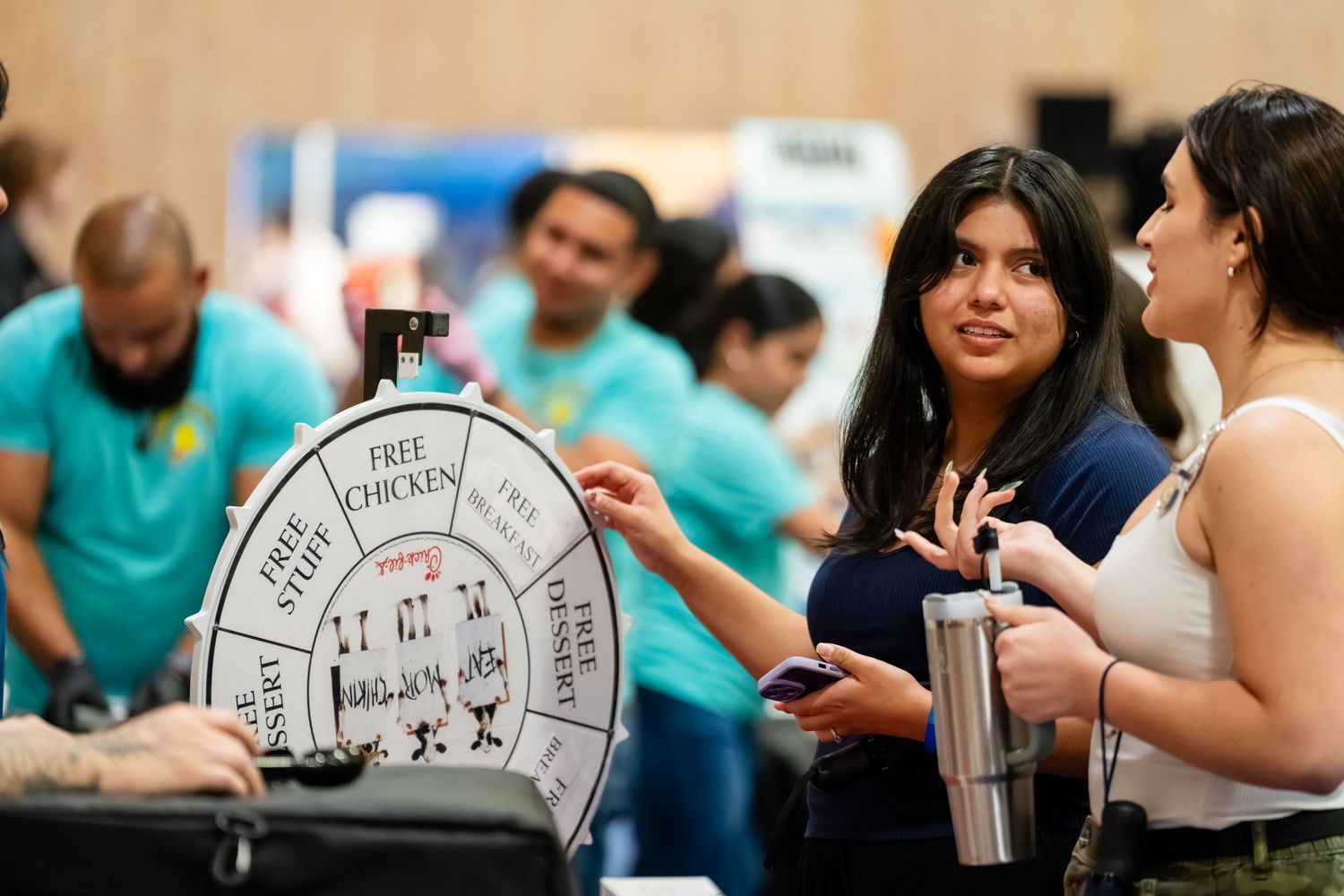 Two women chat beside a prize spin wheel at an indoor event, with people in teal shirts visible in the background.
