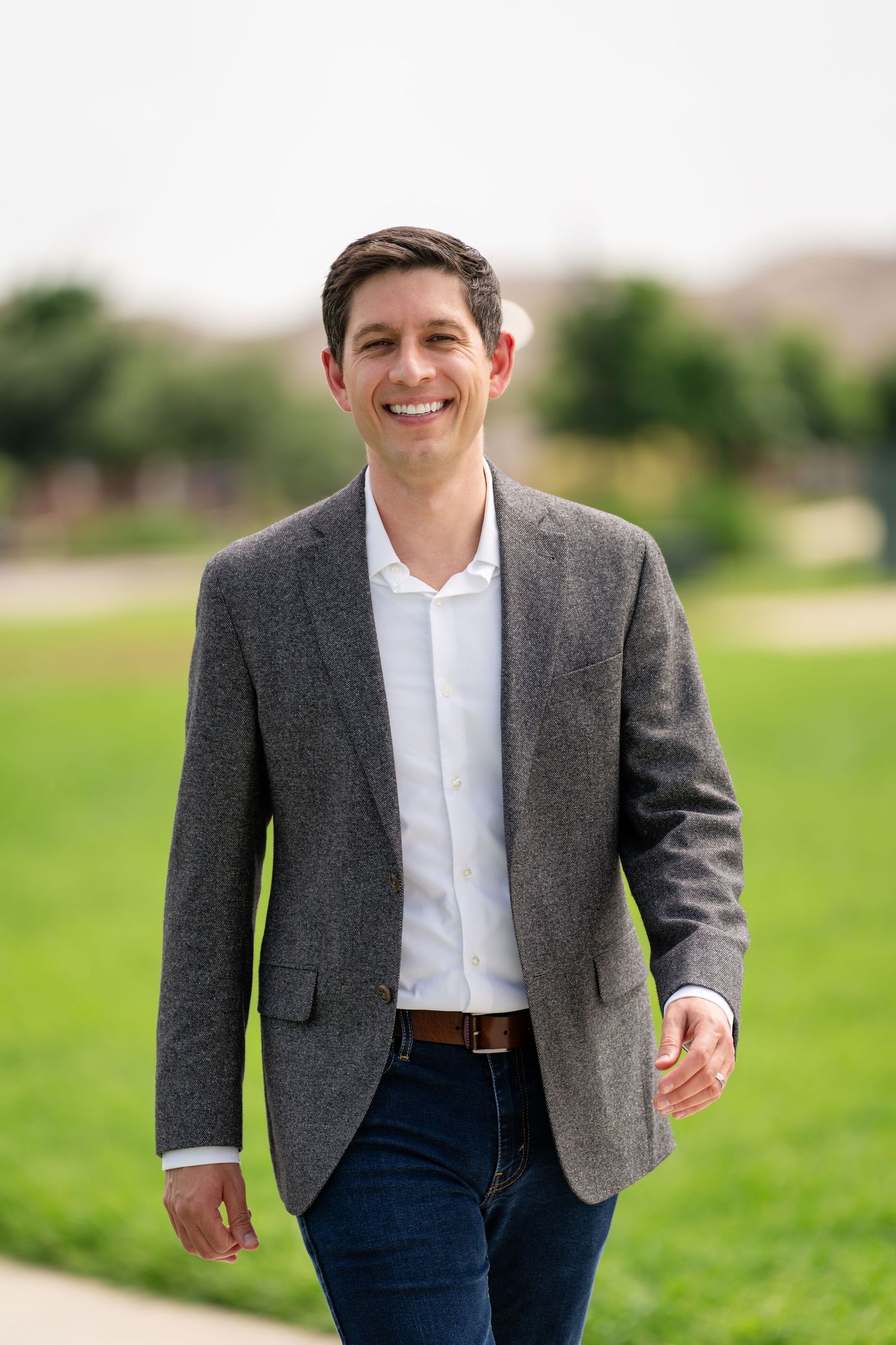 Smiling man in gray blazer and jeans walking outdoors on a sunny day in a green park setting.