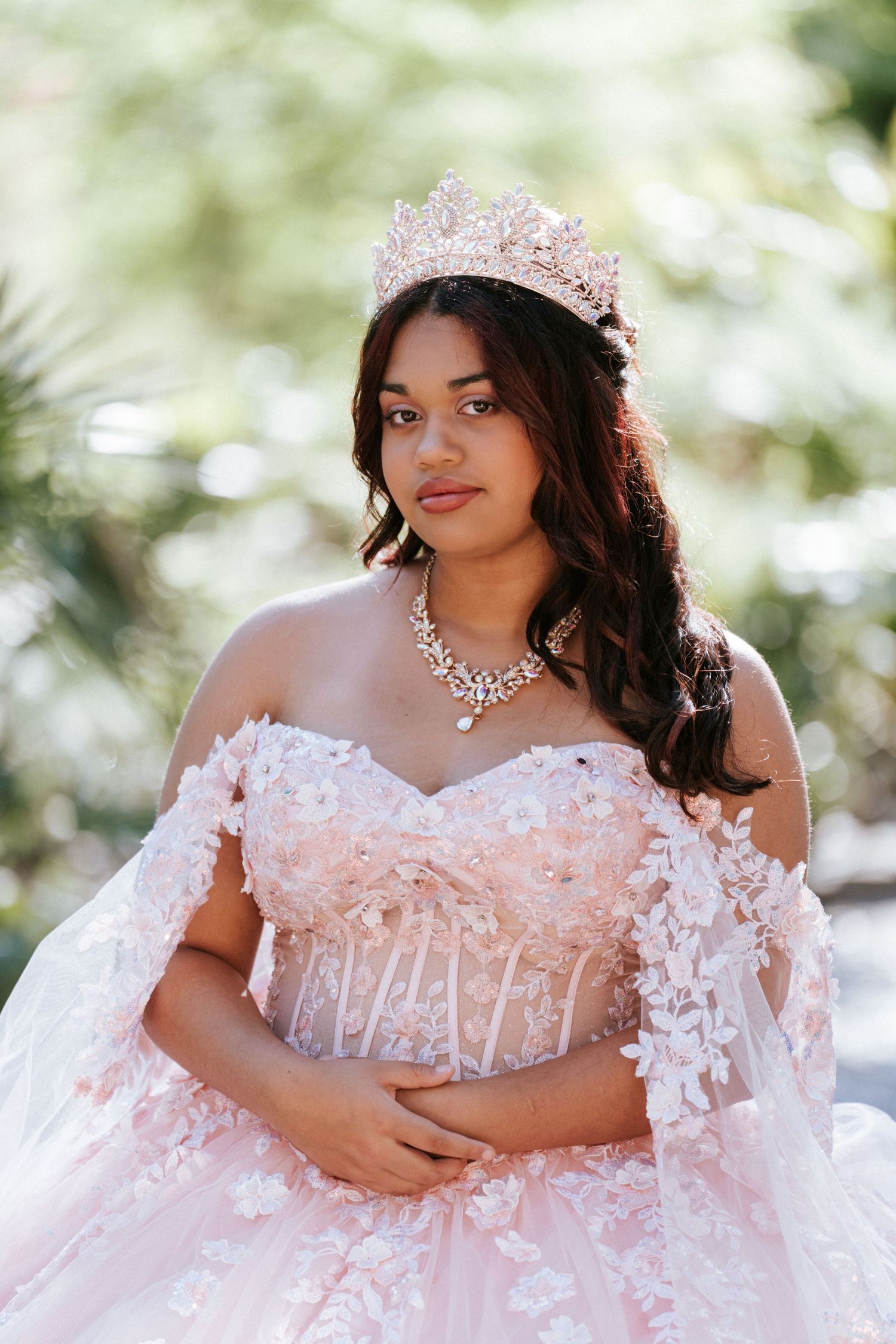 Young woman in pink floral ball gown with crown and crystal necklace poses outdoors for quinceañera portrait.