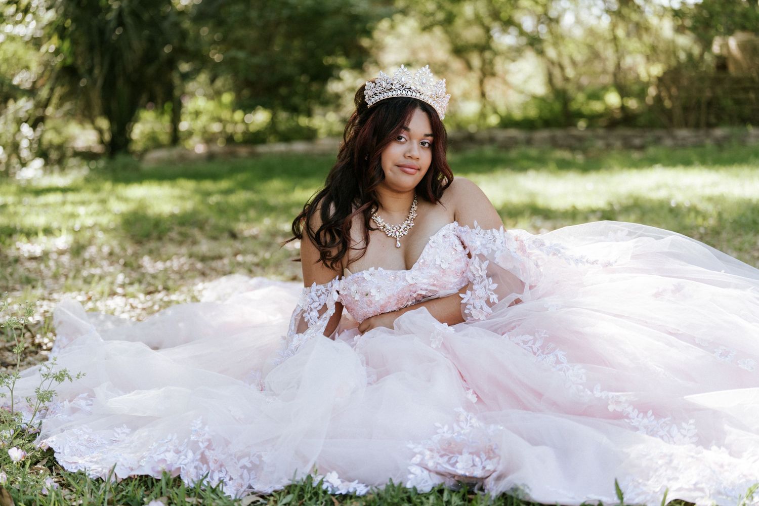 Young woman in pink quinceañera ball gown with lace details and tiara poses gracefully outdoors on green grass.