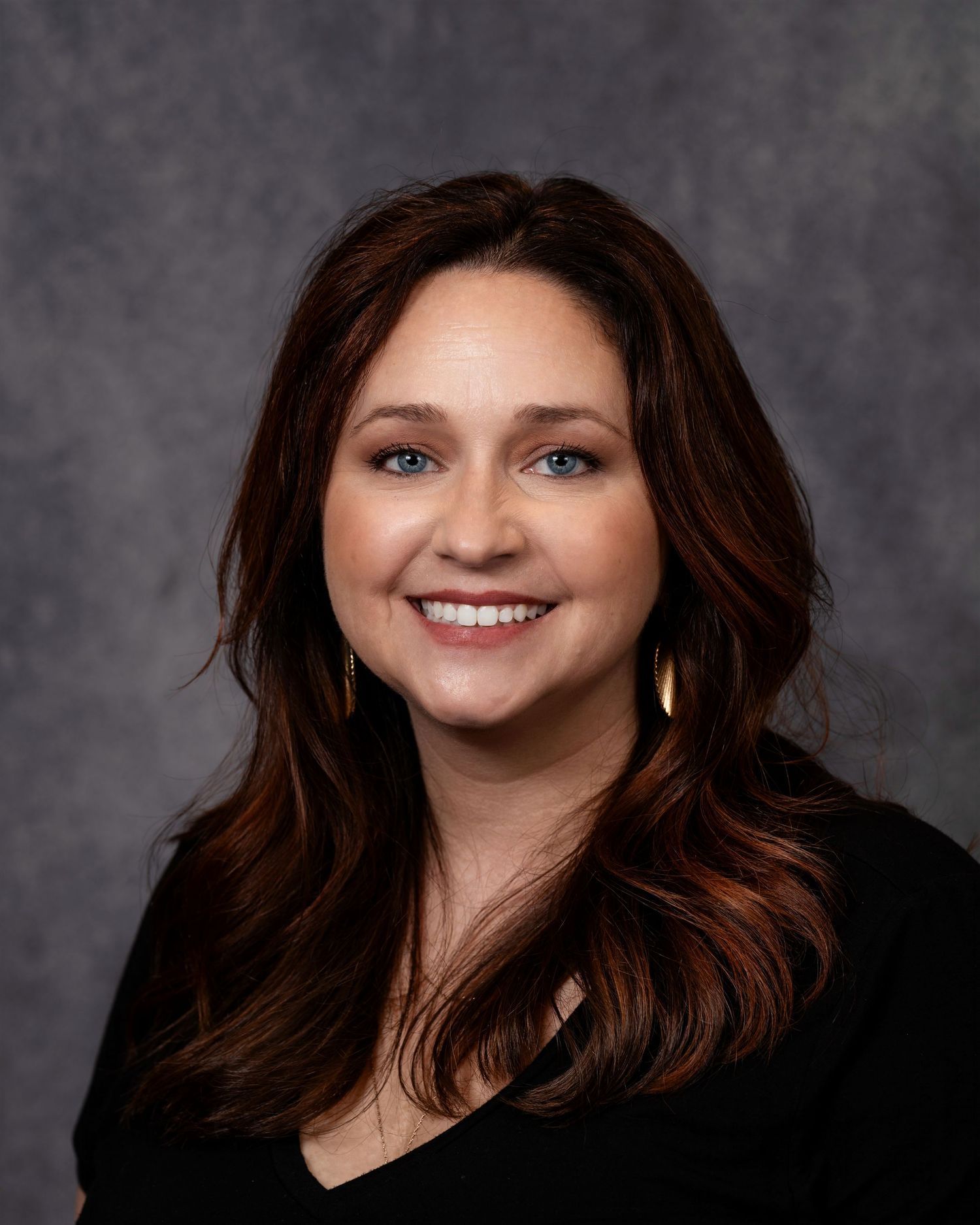 Professional headshot of a smiling woman with long dark auburn hair, blue eyes, and gold earrings against a gray background.